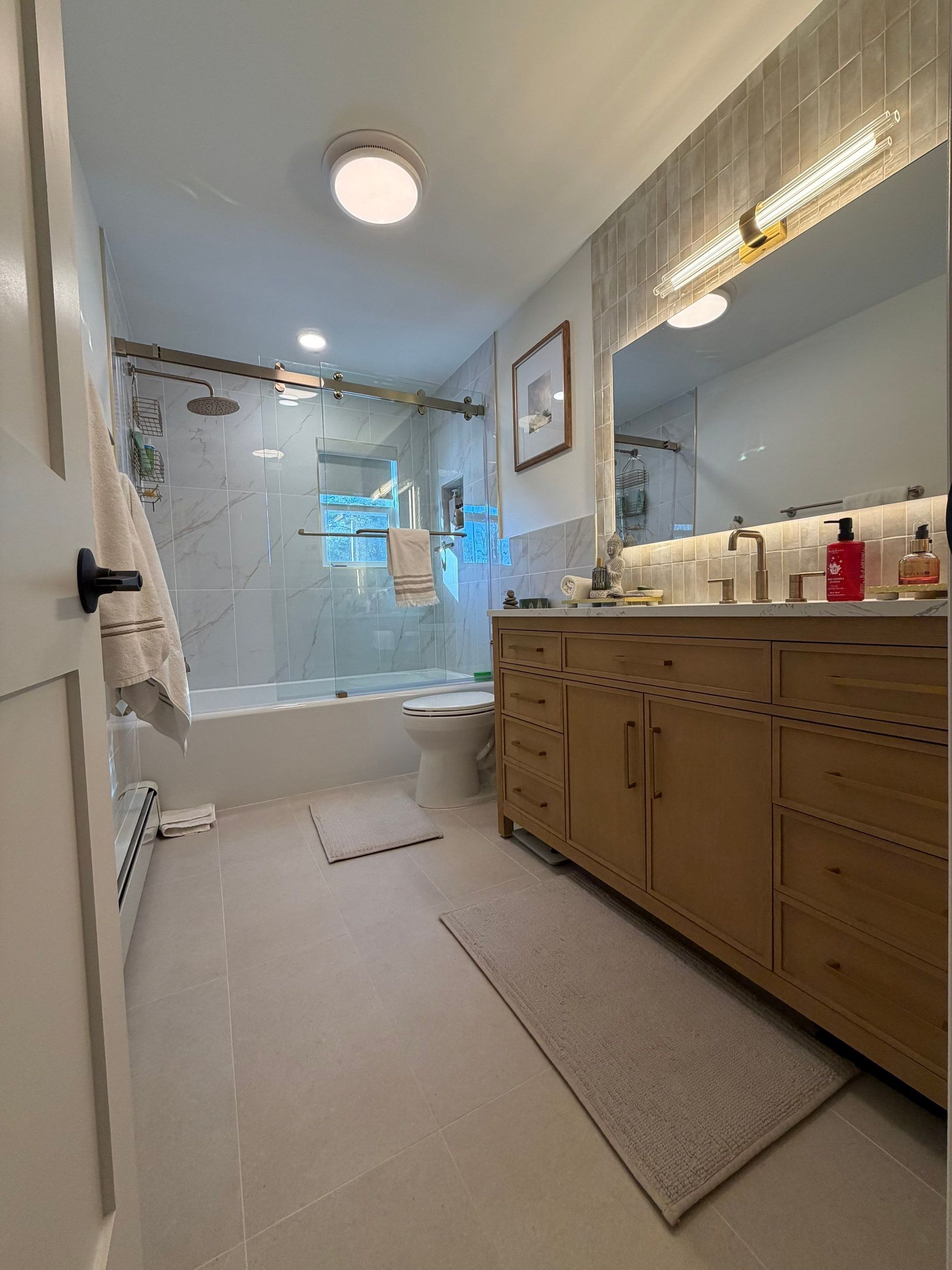Bathroom with light-colored tile, wooden vanity, shower, and a large mirror with a light fixture above it.
