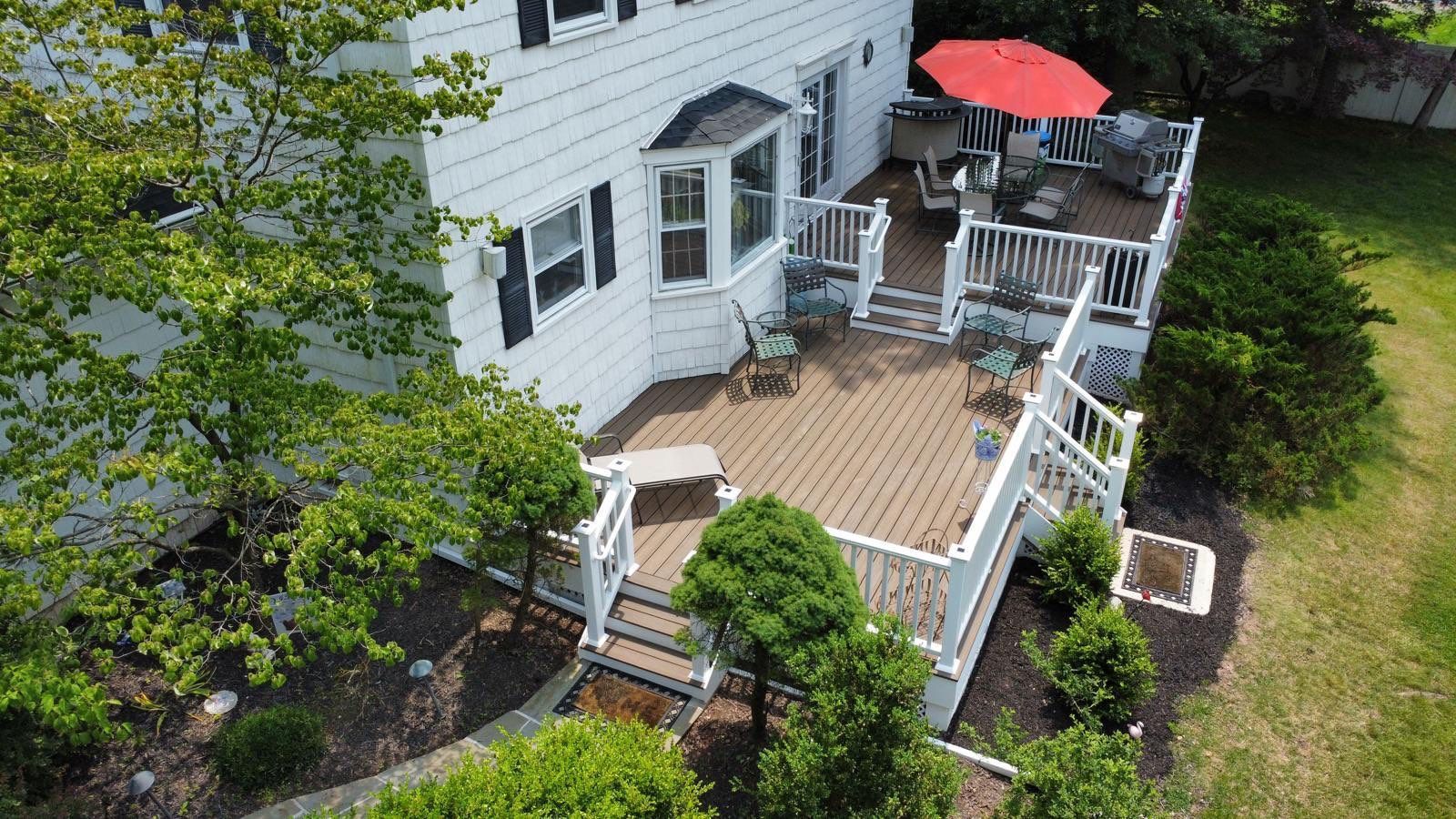 Aerial view of a white house with a multi-level brown deck. Red umbrella, seating, and greenery visible.