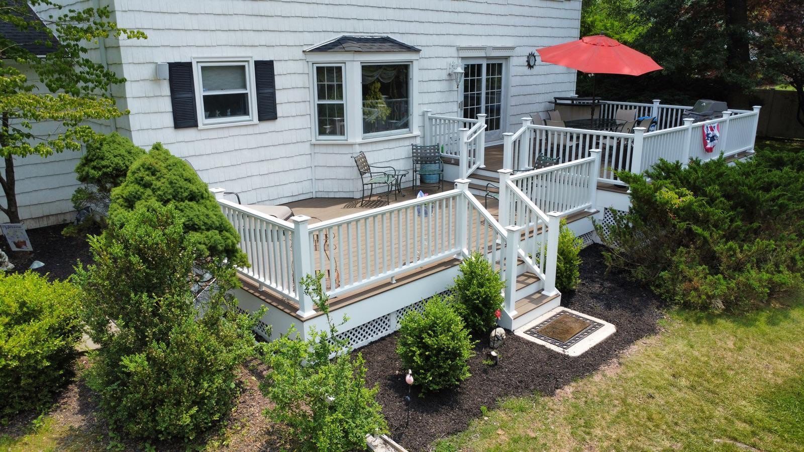 White deck with red umbrella, attached to a white house with dark shutters. Lush greenery surrounds.