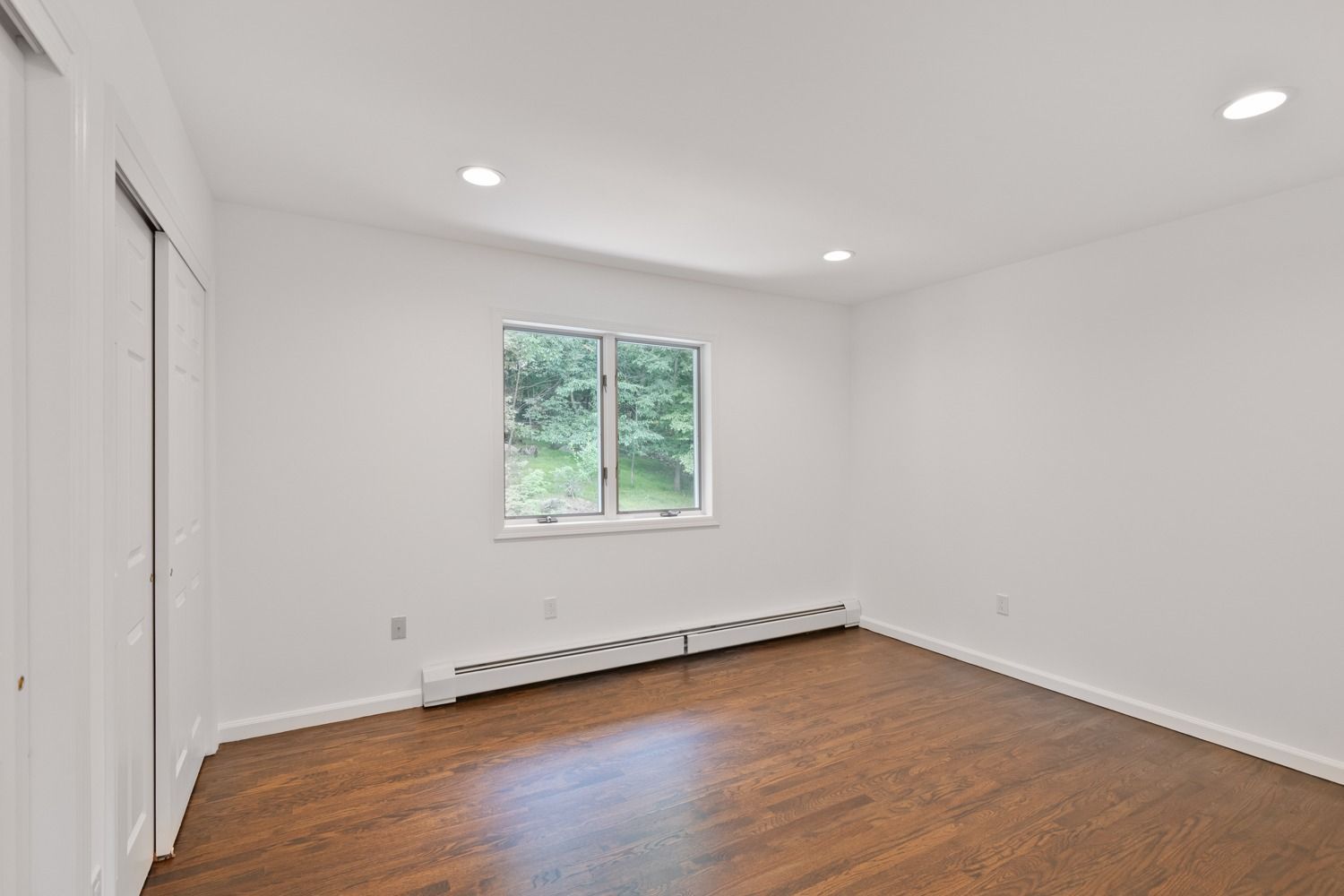 Empty bedroom with hardwood floor, window, and closet doors. White walls and recessed lighting.