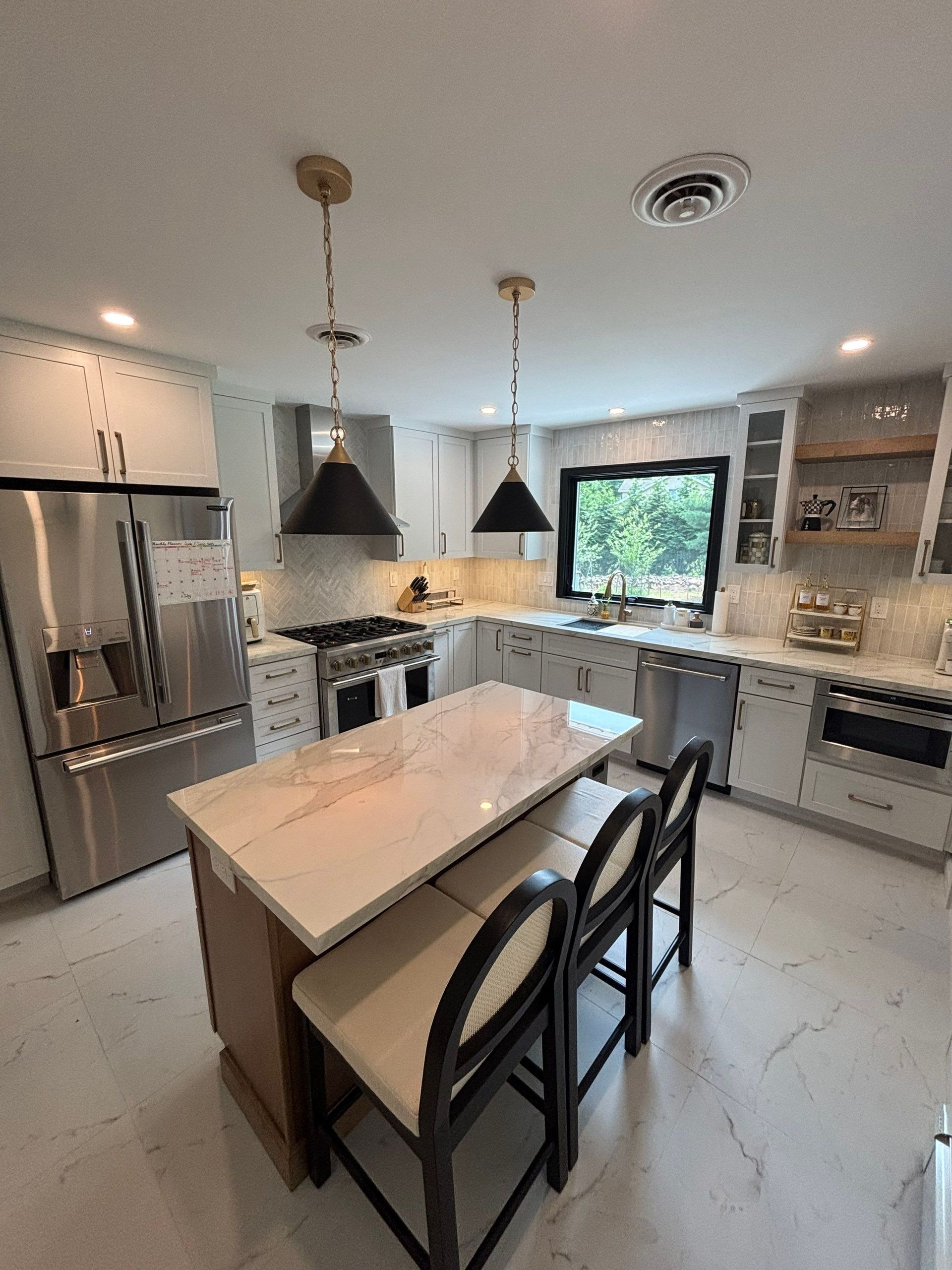 Modern kitchen with island and stools. White cabinets, marble countertops, stainless steel appliances, and black pendant lights.