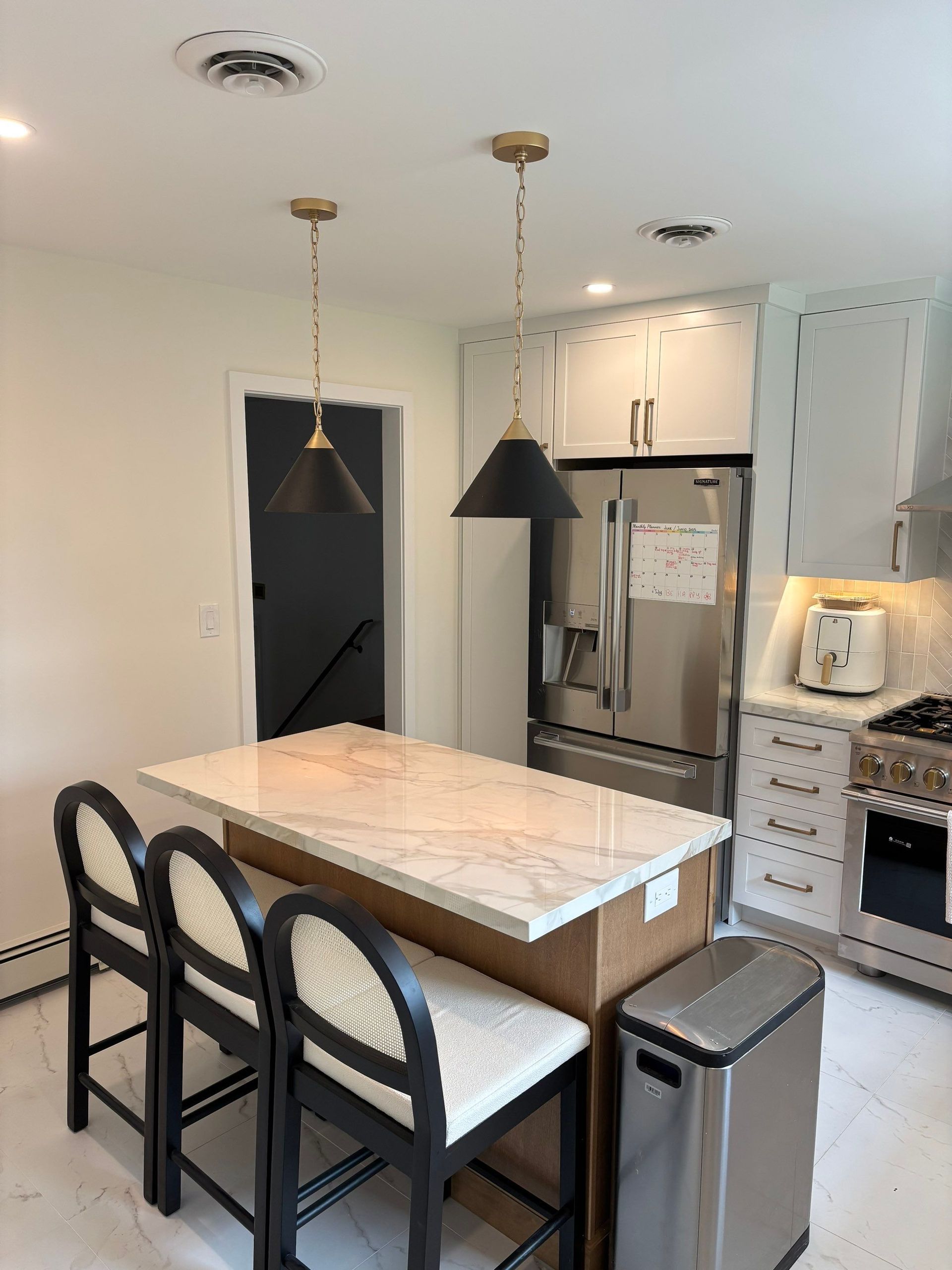 Kitchen with marble countertop island, two black pendant lights, and bar stools. Stainless steel refrigerator and oven.