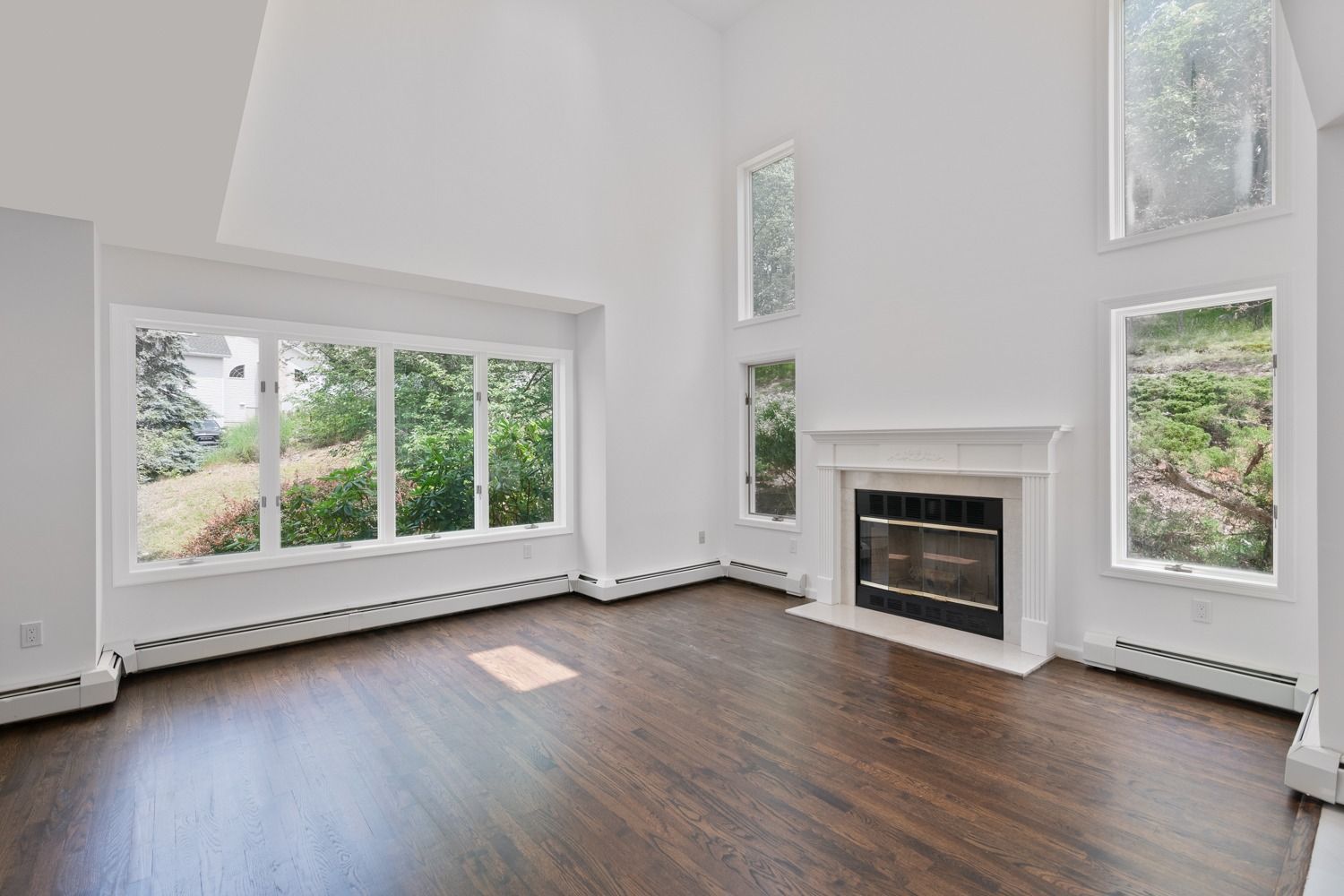 Spacious, empty living room with dark wood floors, white walls, fireplace, and large windows overlooking greenery.