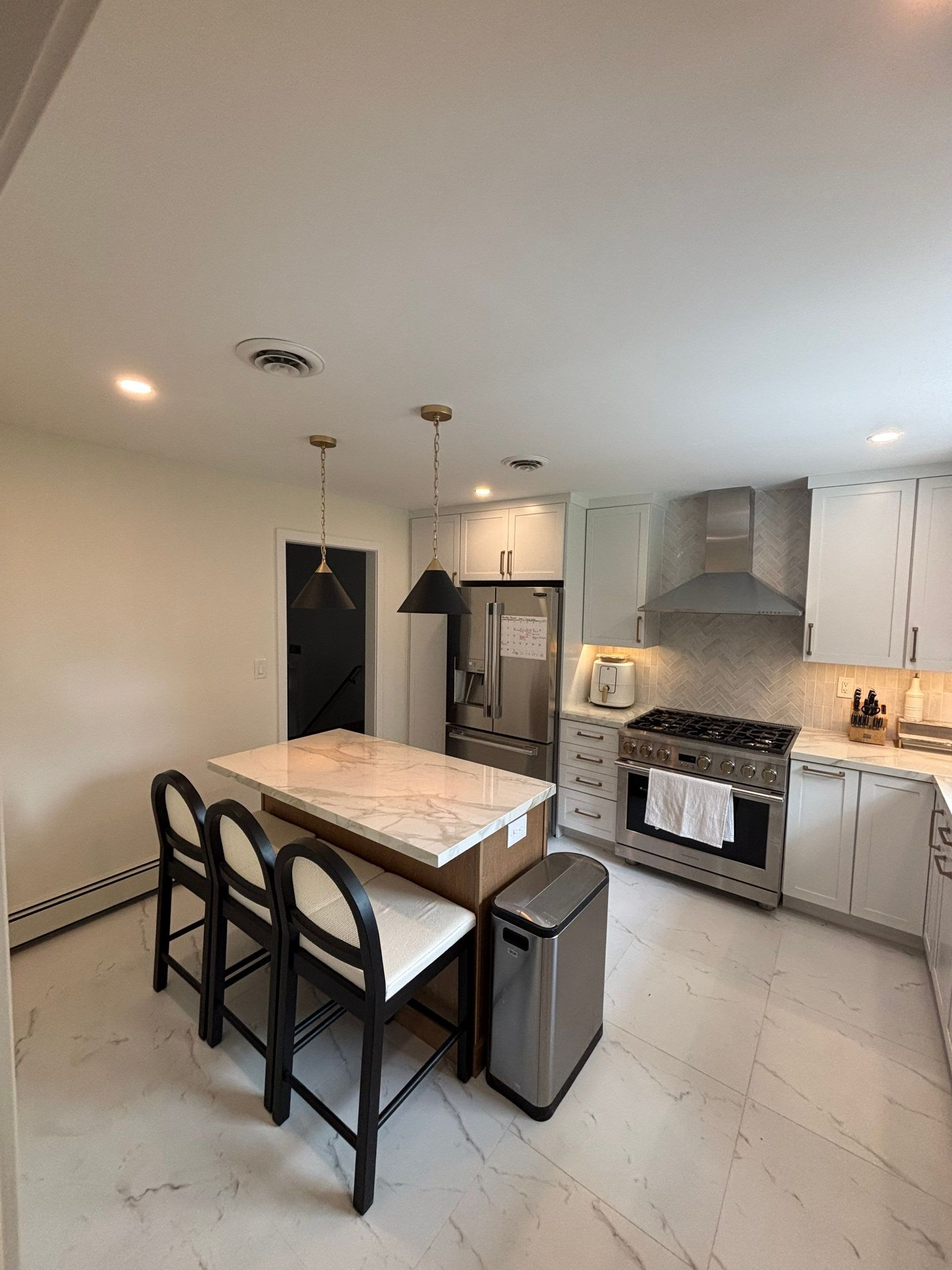 Modern kitchen with white cabinets, island with stools, stainless steel appliances, and a marble floor.