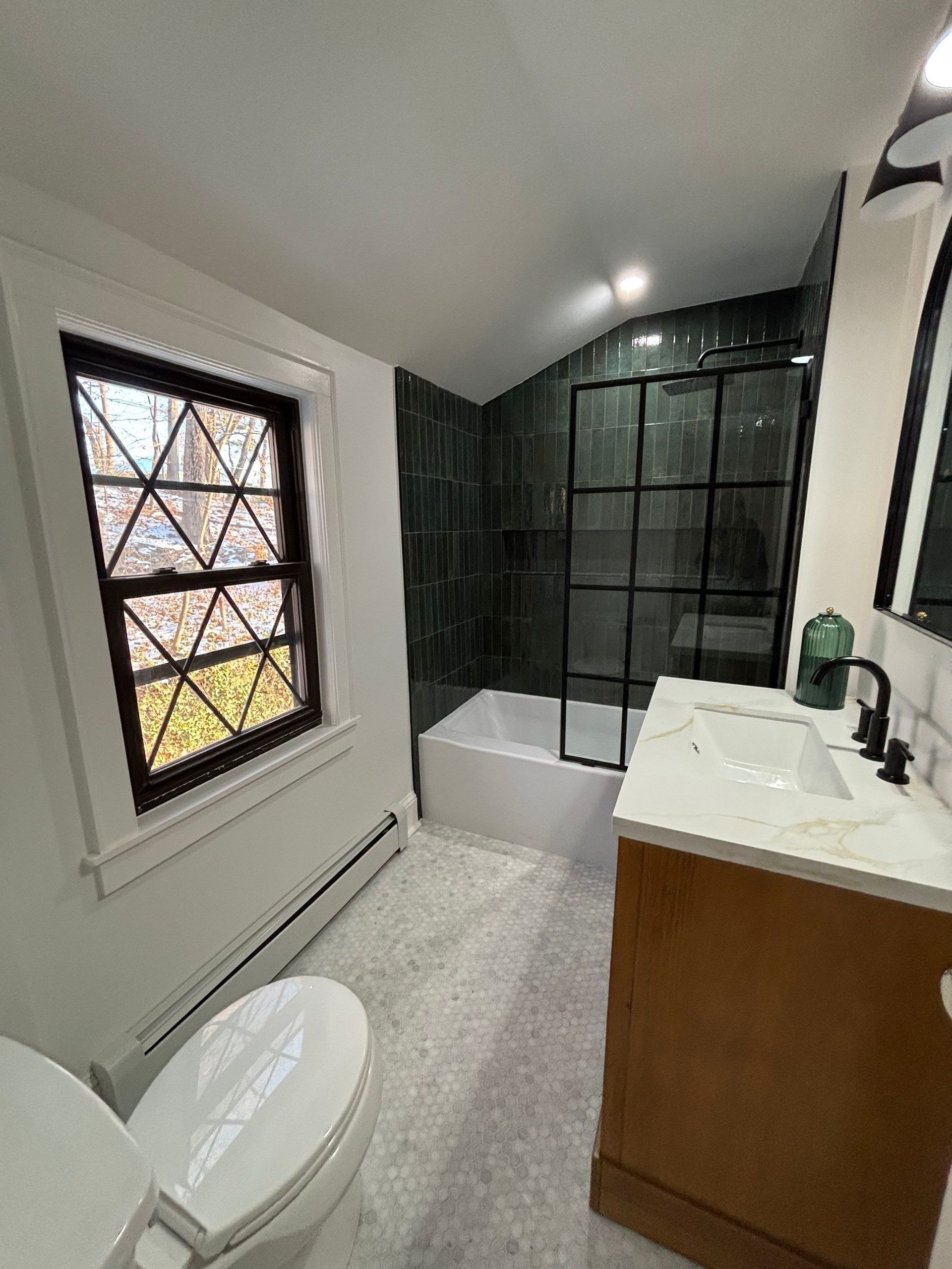 Bathroom with a tub, glass shower door, vanity, toilet, and a window. Light-colored floor and walls, dark tile shower surround.