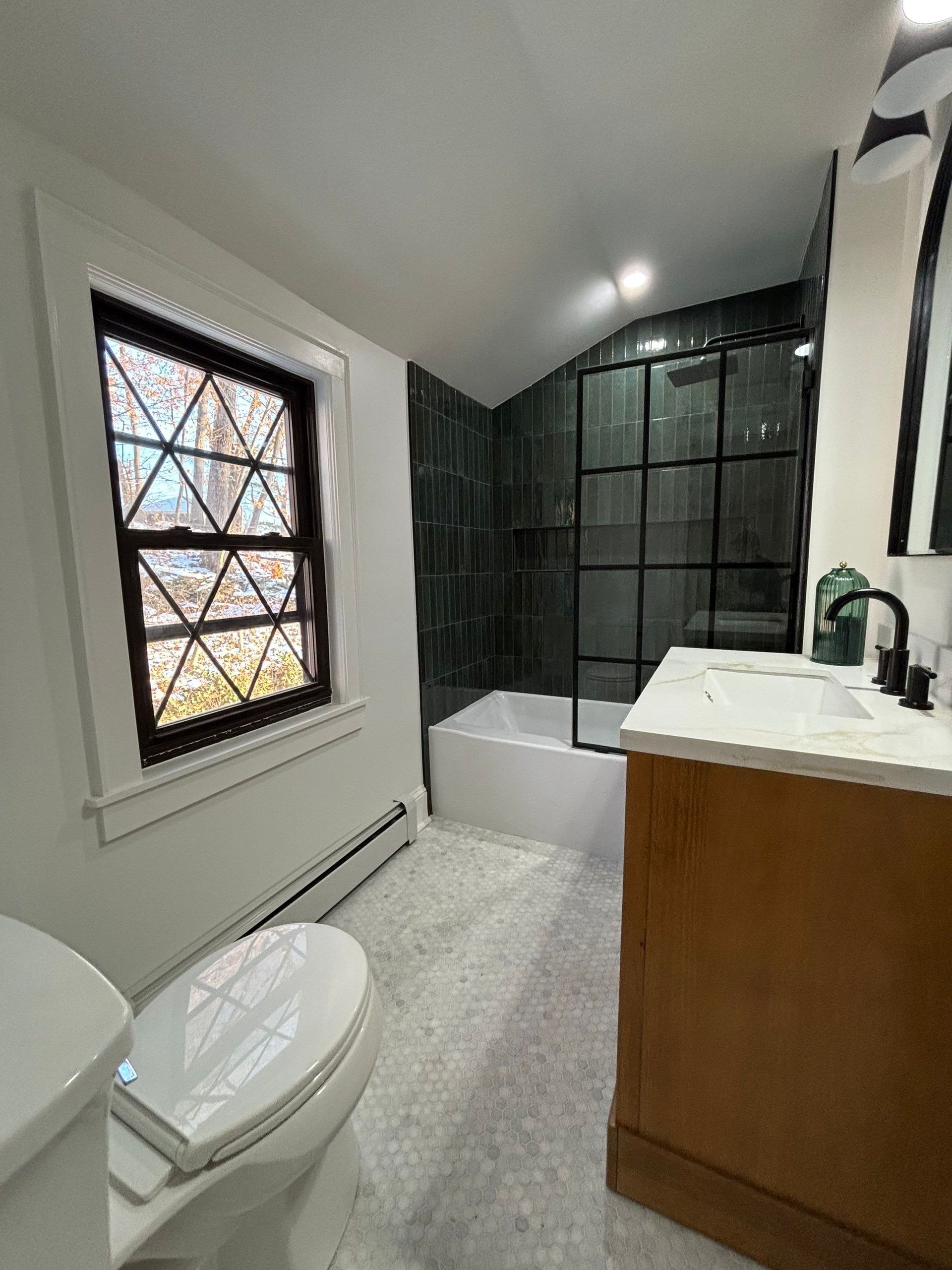 Bathroom with black-framed shower, tile, vanity, and window. Neutral tones dominate.