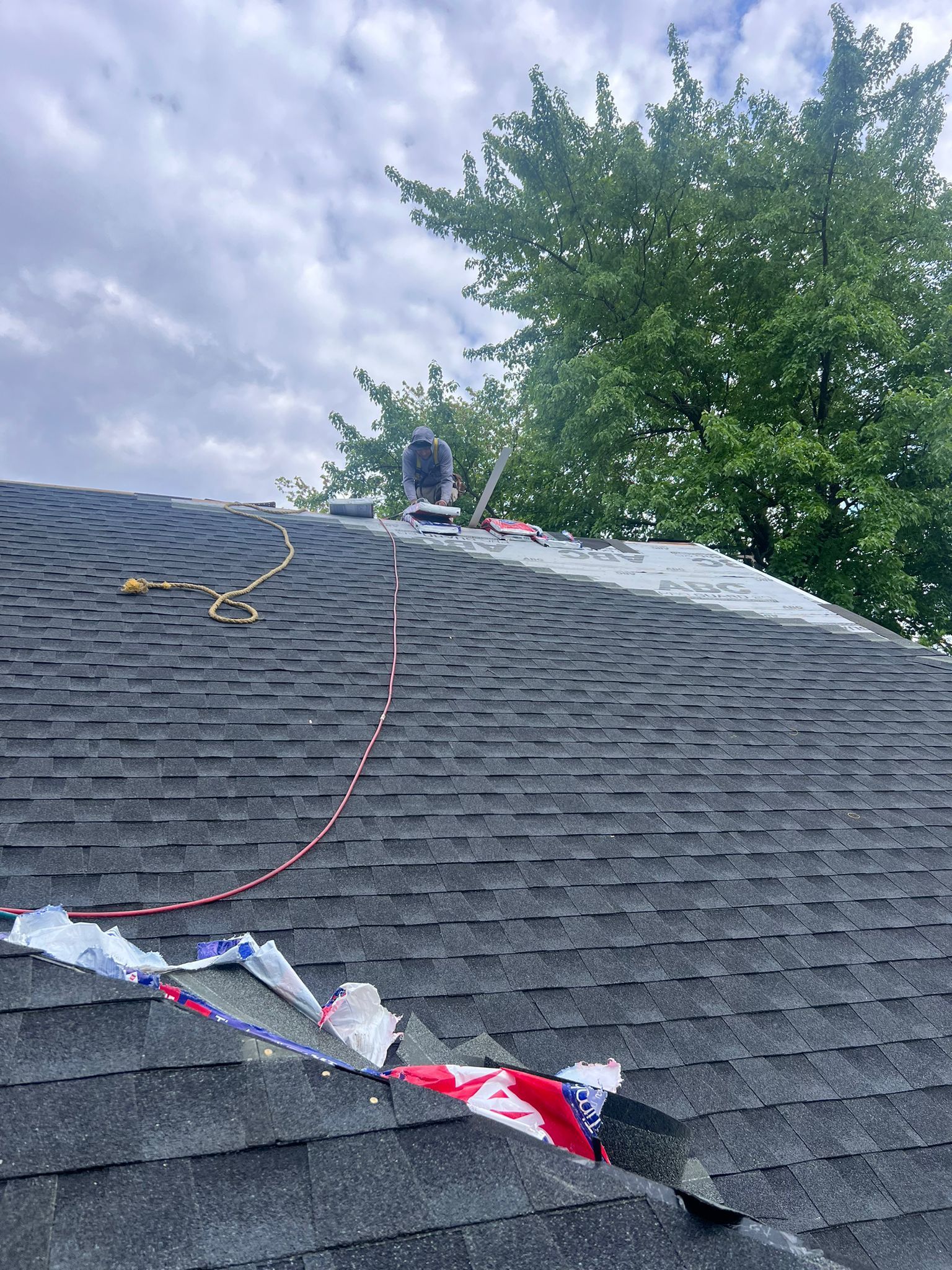 Roofer working on a dark shingle roof with a cloudy sky and green tree in the background. Debris present.