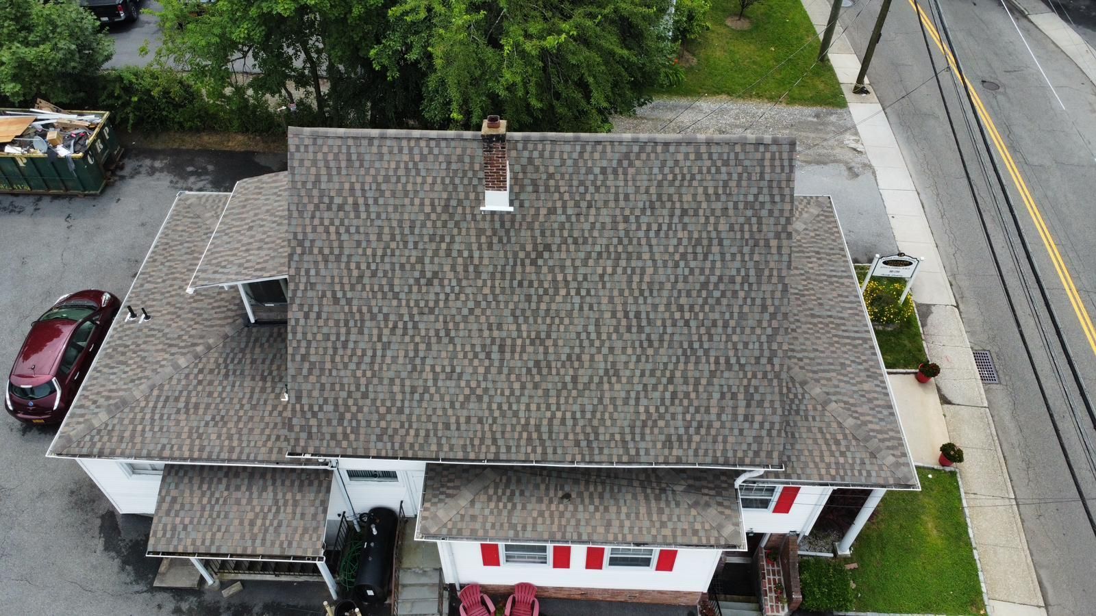 Aerial view of a building with a gray shingled roof, chimney, and red trim, next to a road.