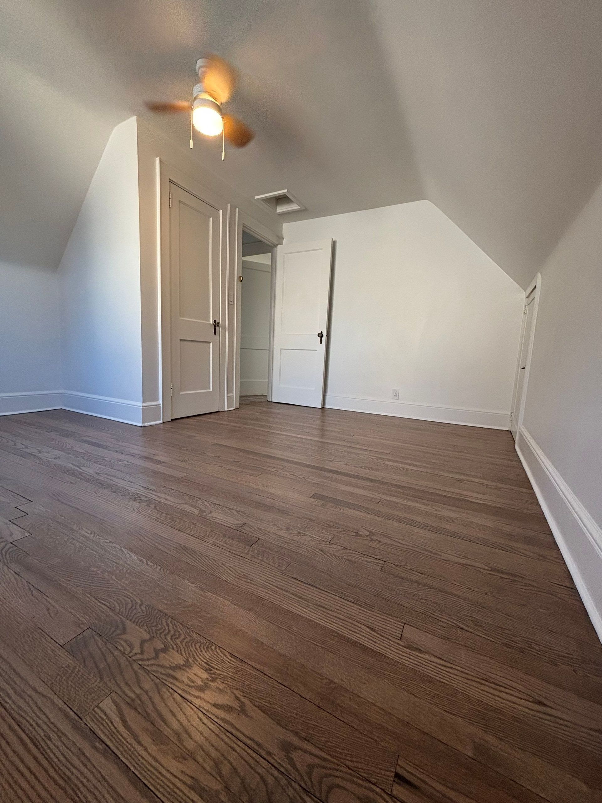 Empty attic room with hardwood floors, white walls, and a ceiling fan.
