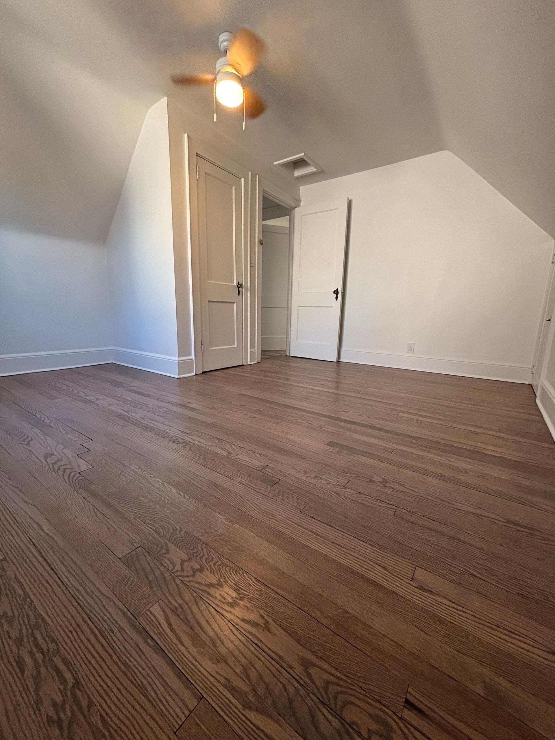 Wooden-floored attic bedroom with sloped white walls, two white doors, and a ceiling fan.
