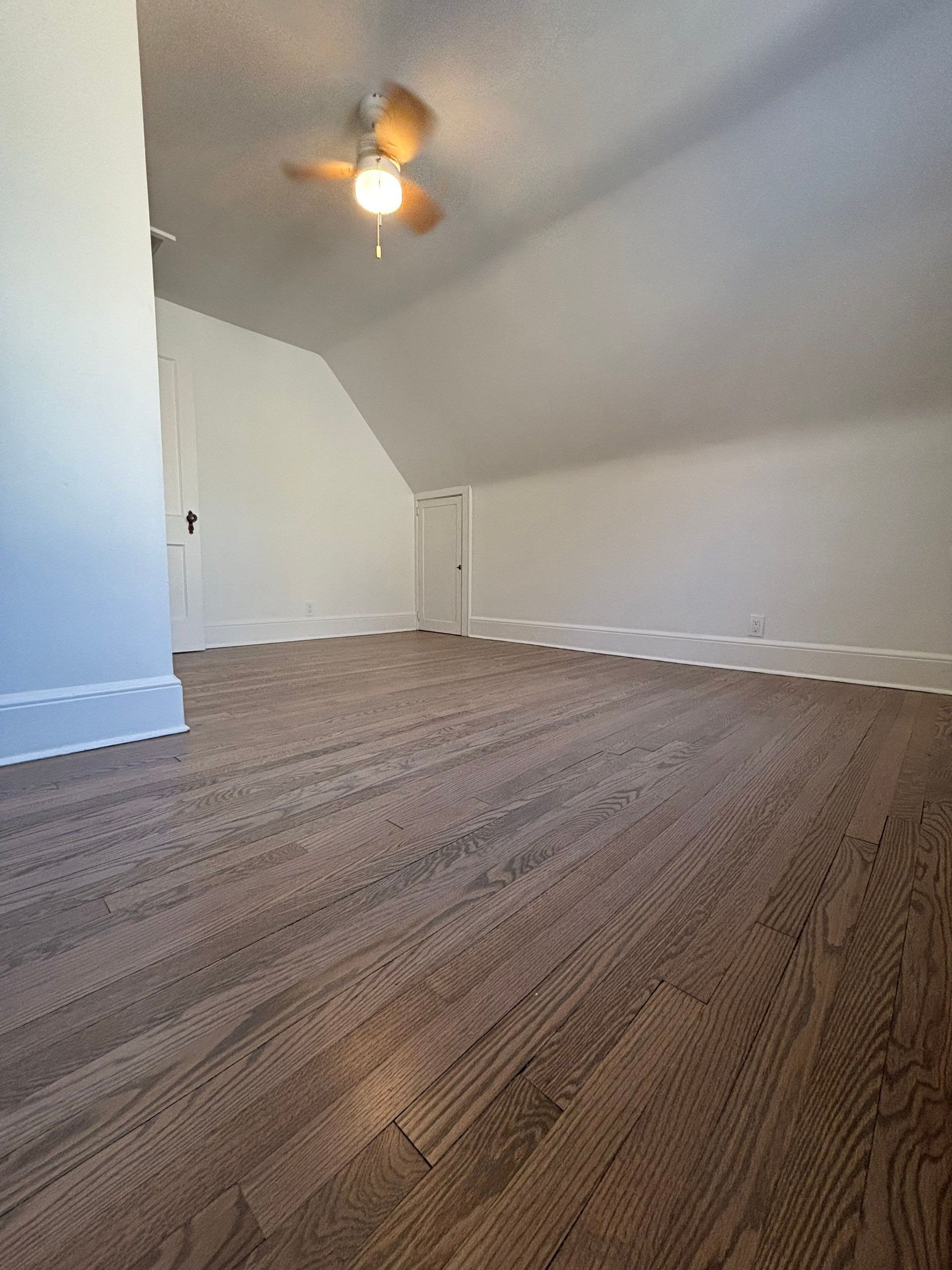 An attic room with angled ceilings, wood flooring, and a ceiling fan.