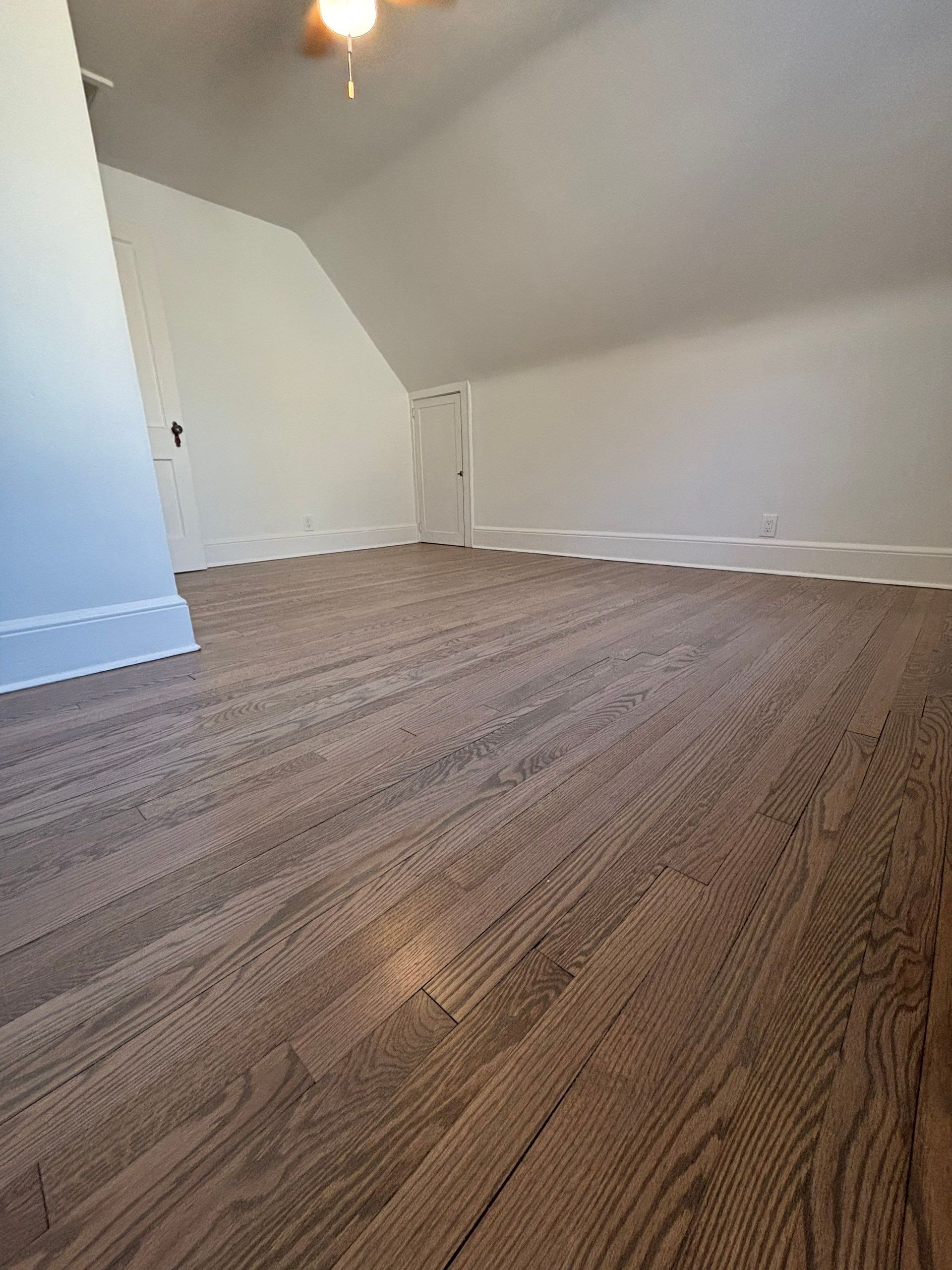 Spacious attic room with angled ceilings, light brown wood floor, and white walls.