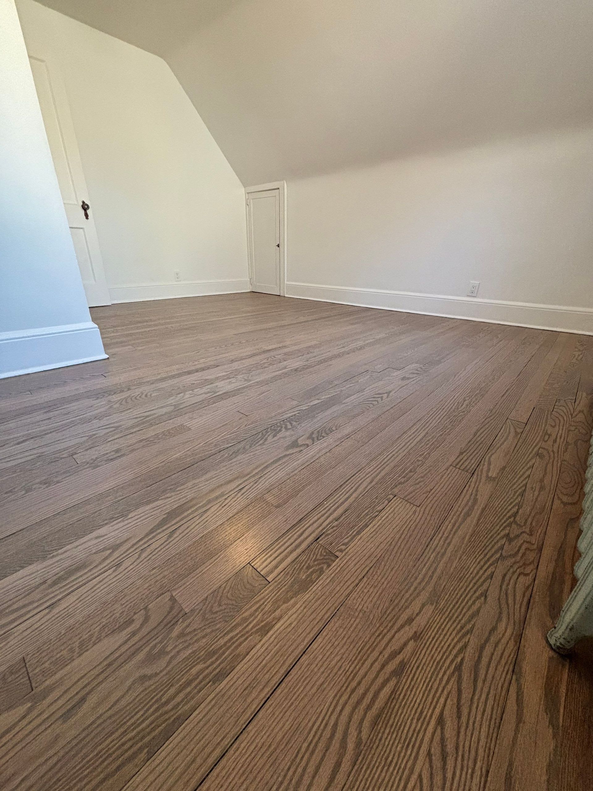 Wooden floor in an attic room with white walls, slanted ceiling, and a small door.