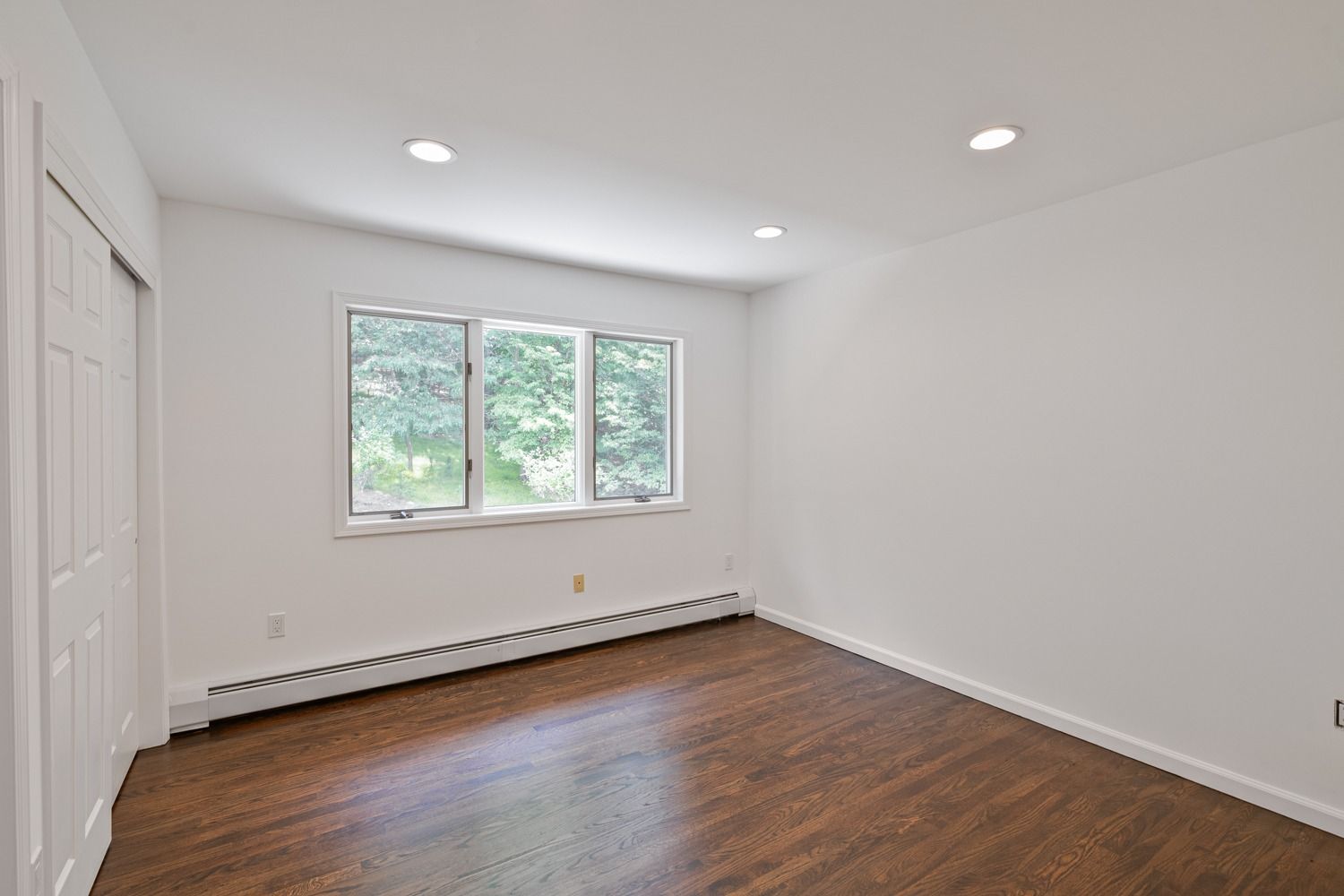 Empty bedroom with hardwood floor, white walls, three windows, and closet.