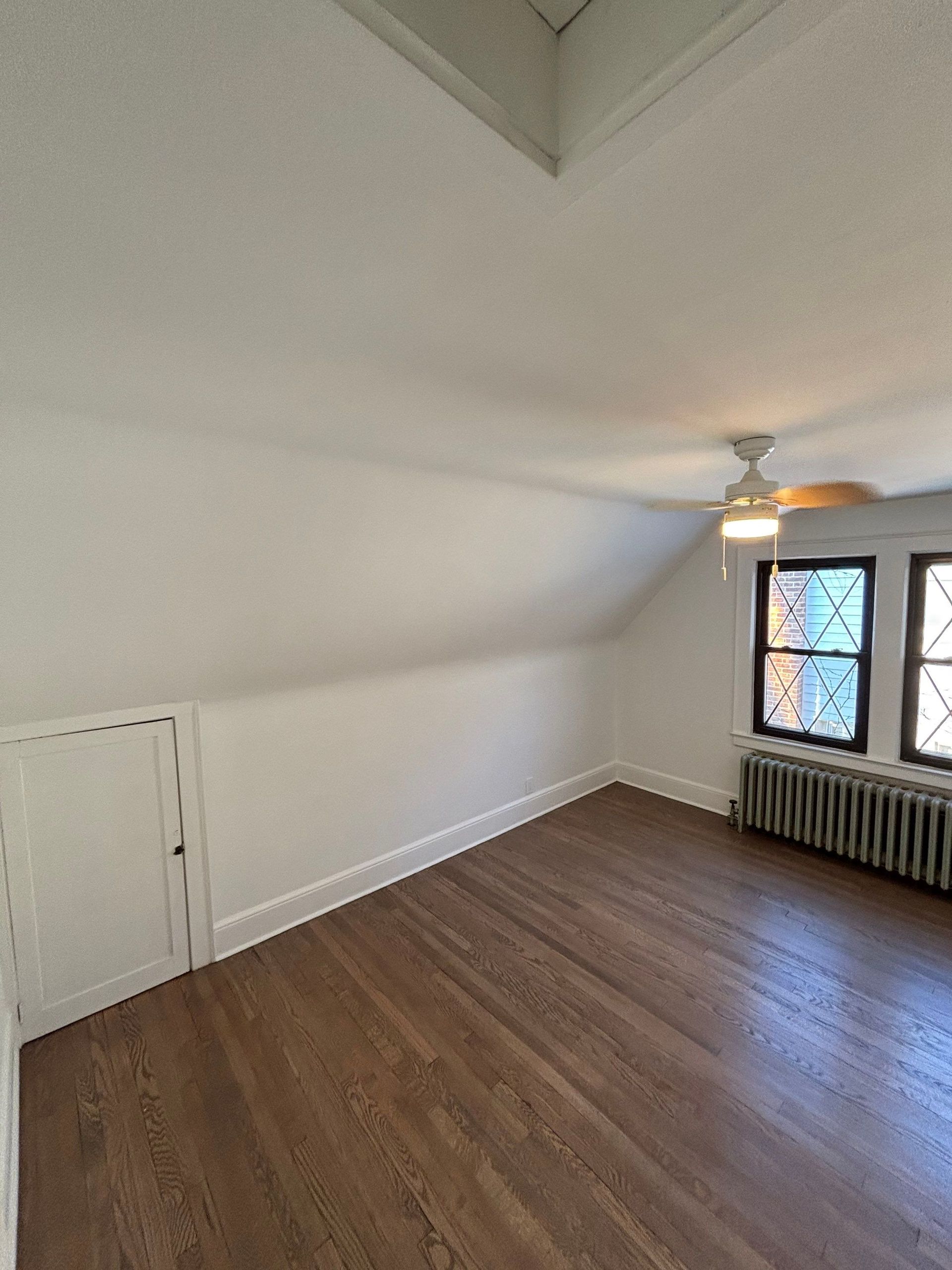Empty room with angled ceiling, wood floor, small window, and storage door.