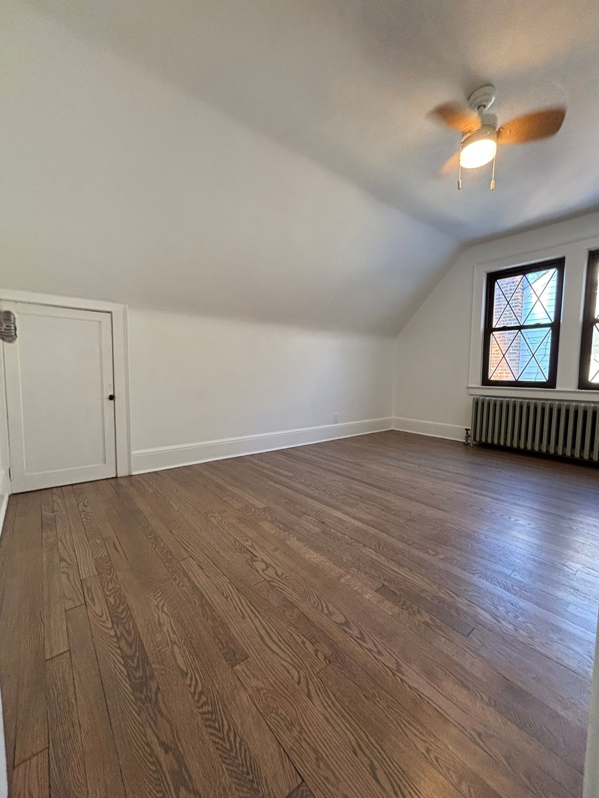 Spacious room with hardwood floors, white walls, and a sloped ceiling with a fan.