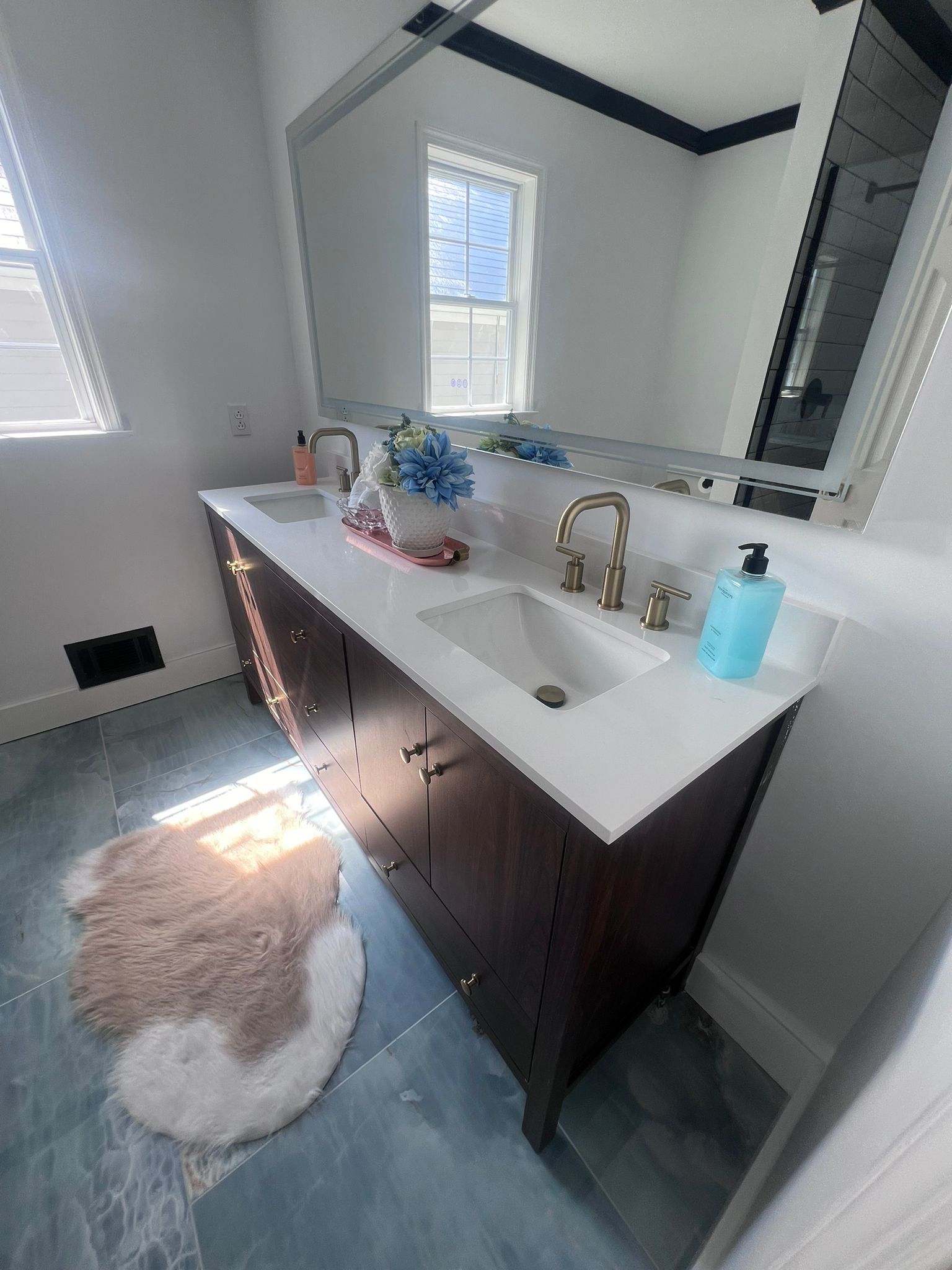 Bathroom with a brown double sink vanity, white countertop, gold fixtures, and a fluffy rug on blue-gray tile.