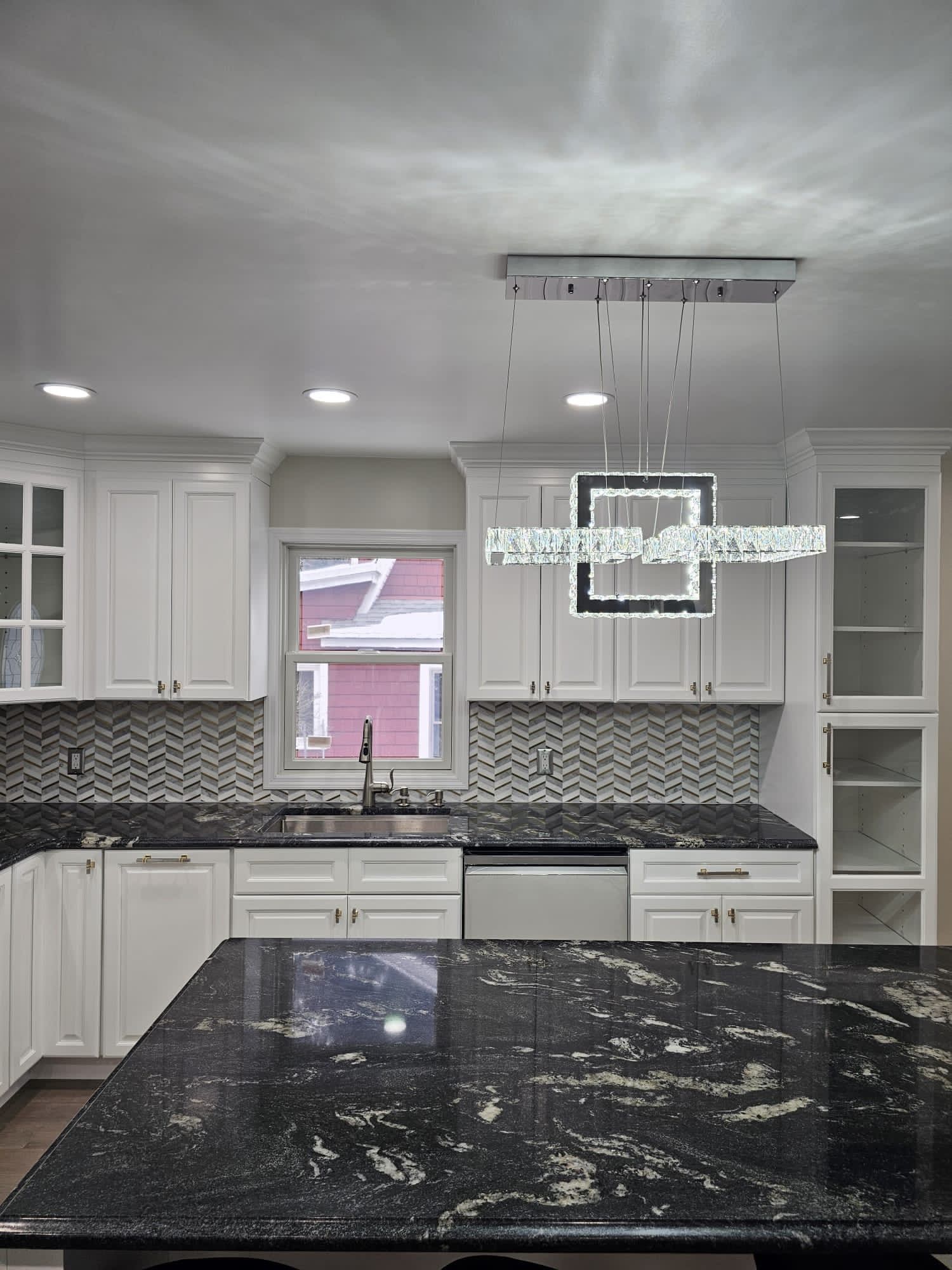 White kitchen with black countertops, chevron backsplash, and modern crystal light fixture.