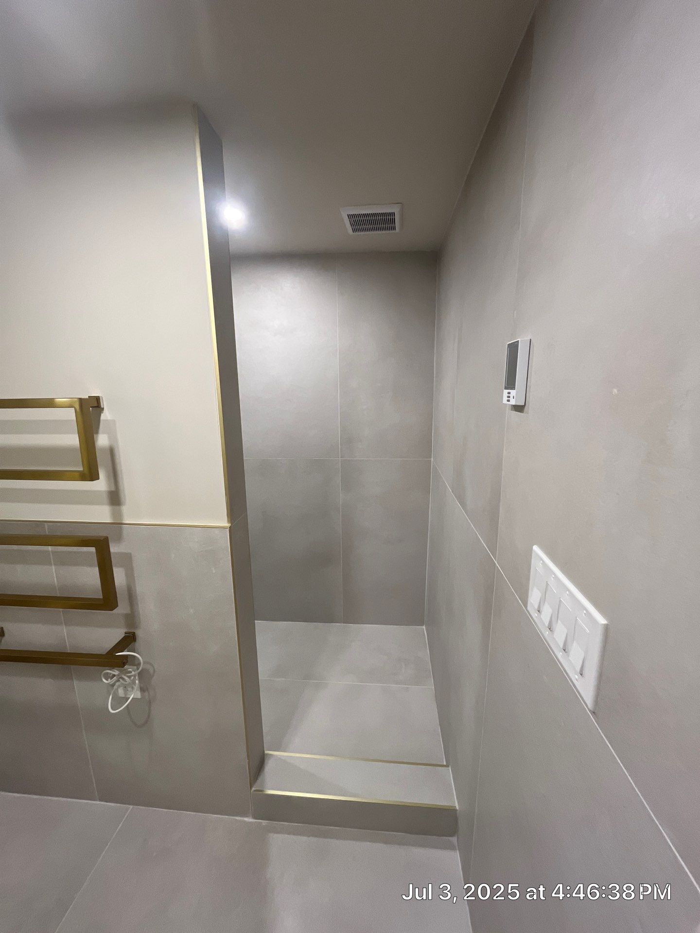 Modern bathroom with grey tiled walls and shower entrance. Gold towel rack on left wall, light switch on the right.