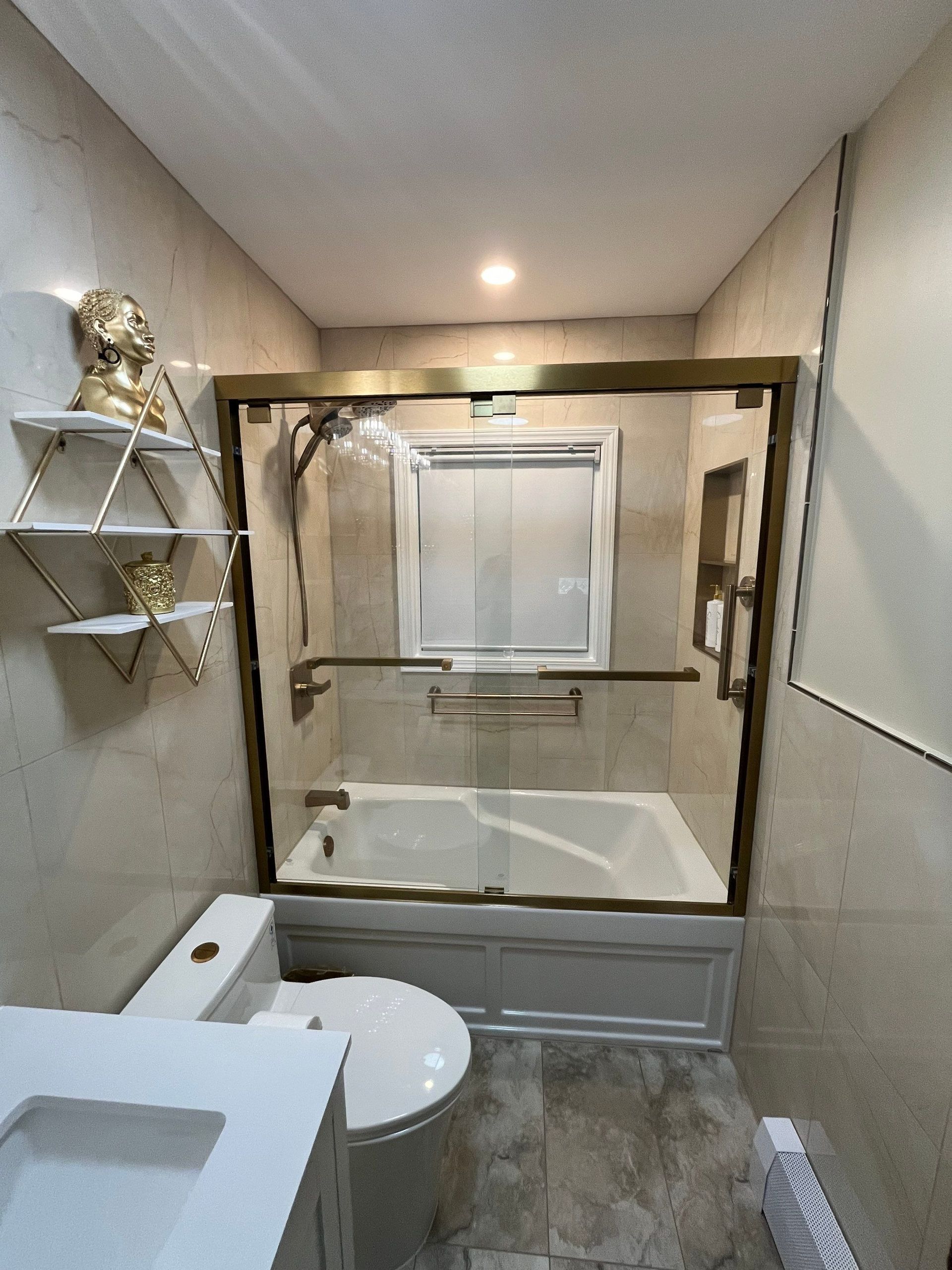 Bathroom with gold-framed glass shower over a bathtub, toilet, shelves, and a window.
