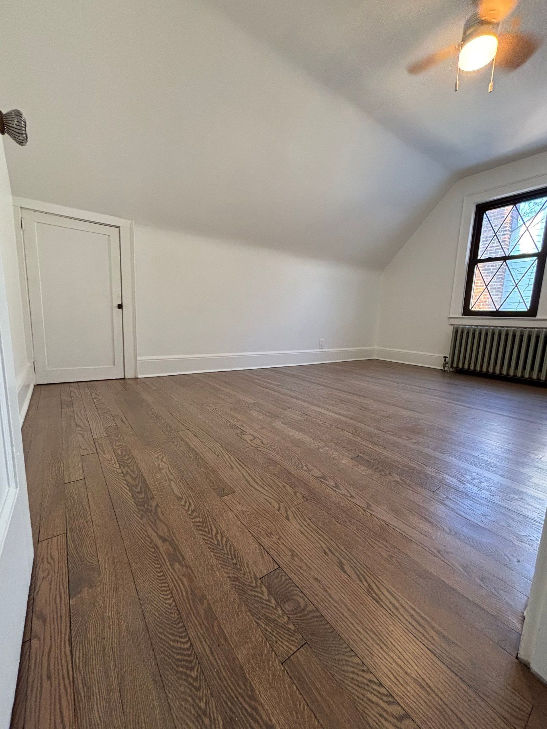 Empty attic room with hardwood floors, white walls, a door, and a window with a radiator.