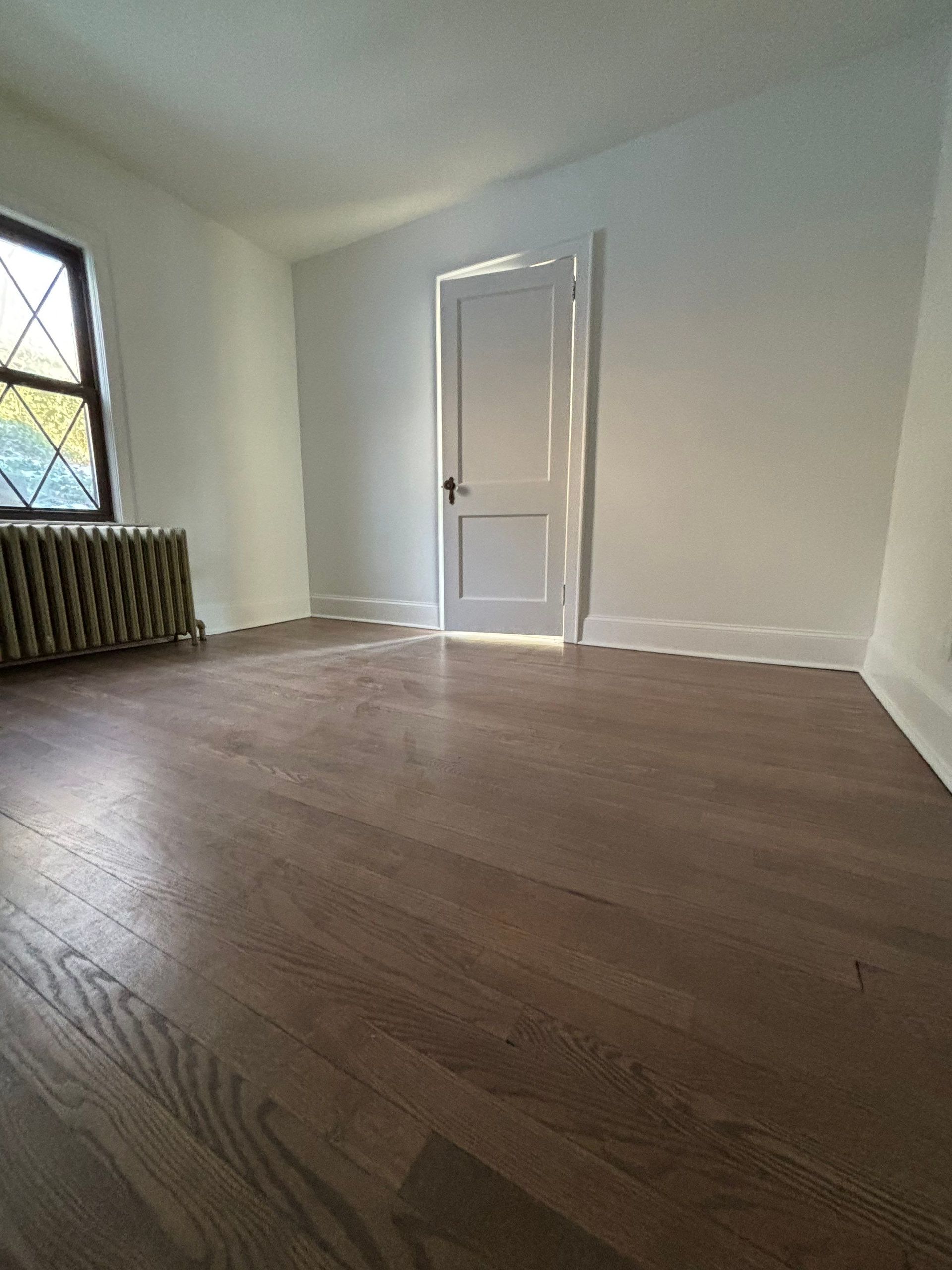 Empty room with hardwood floor, white walls, door, radiator, and a window.