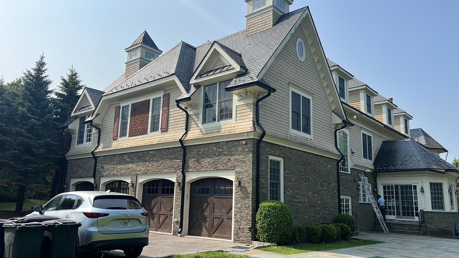 Large house with stone and beige exterior, brown garage doors, and a silver car parked out front on a sunny day.