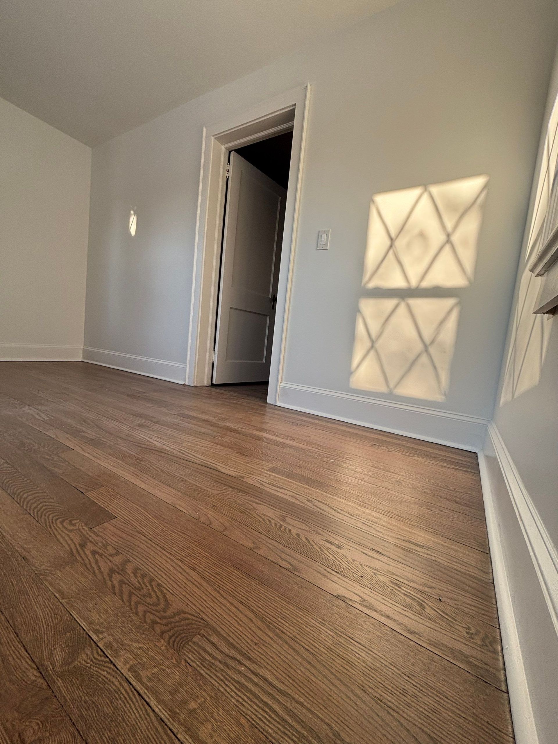 Empty room with hardwood floors, open doorway, and light shadow from a diamond-patterned window.
