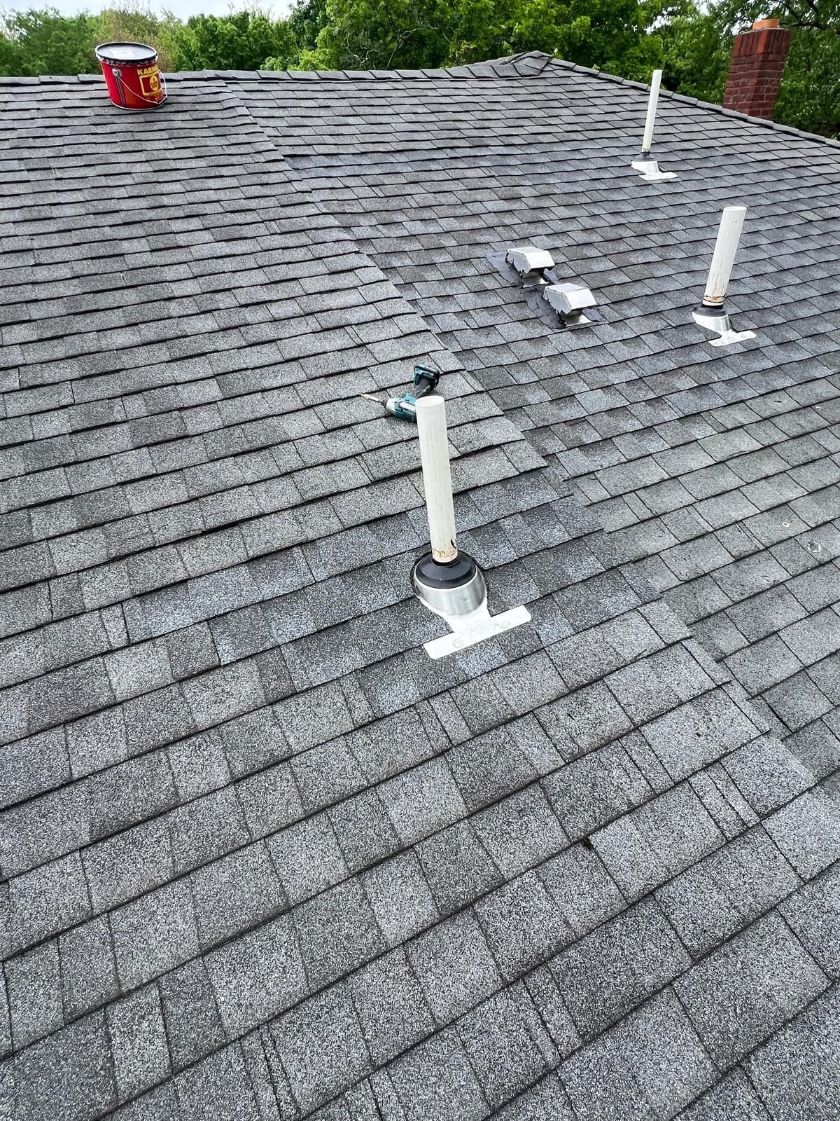 Gray shingle roof with several vents. A red bucket sits on the roof. Green trees in the background.