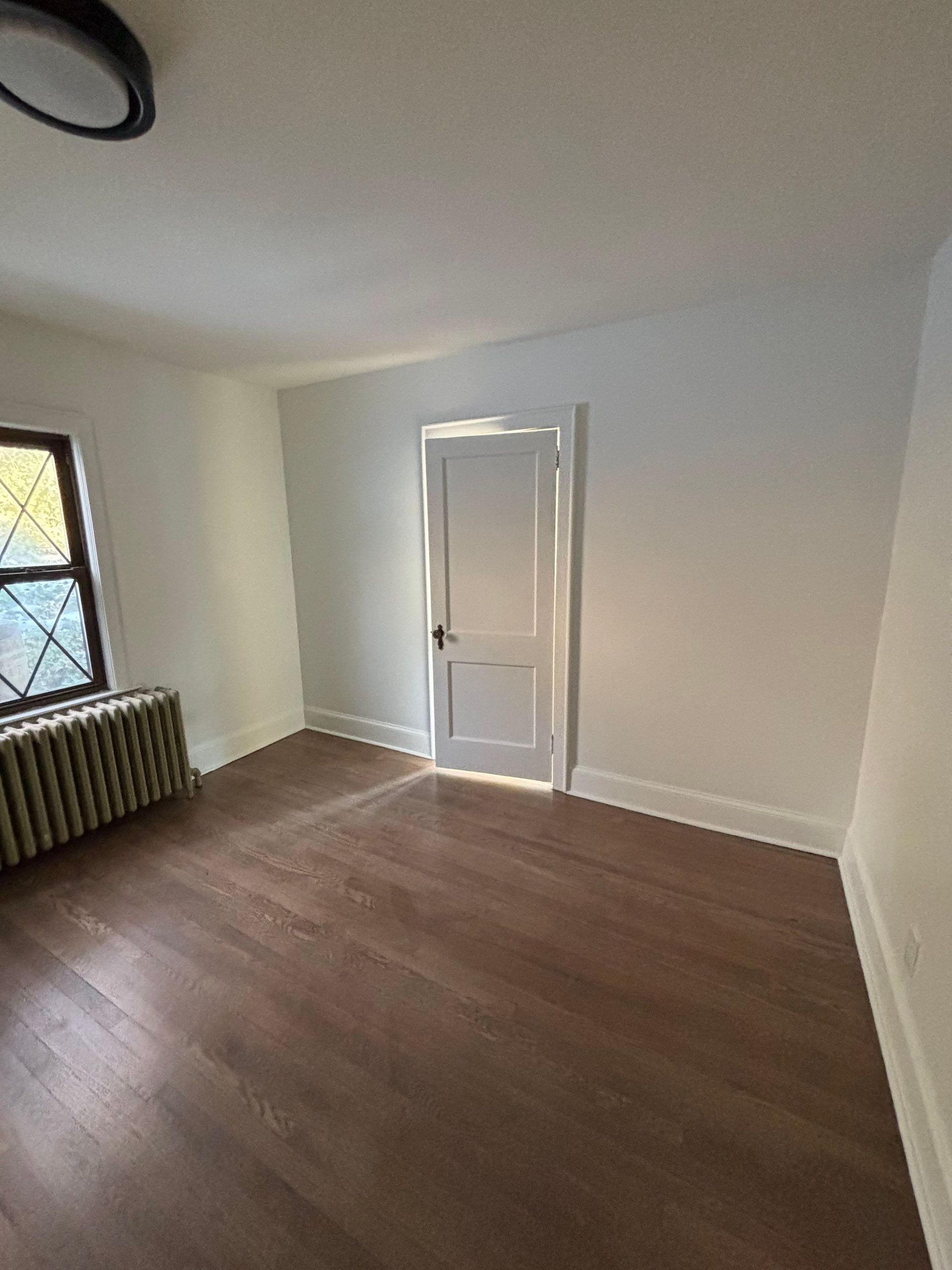 Empty room with wood floor, white walls, door, radiator, and window.
