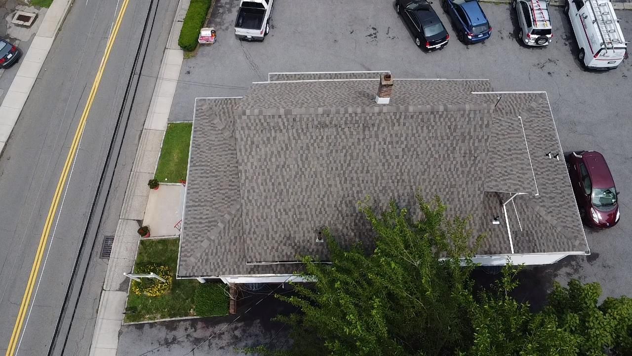 Overhead view of a building with a gray roof, next to a parking lot with cars and a road.
