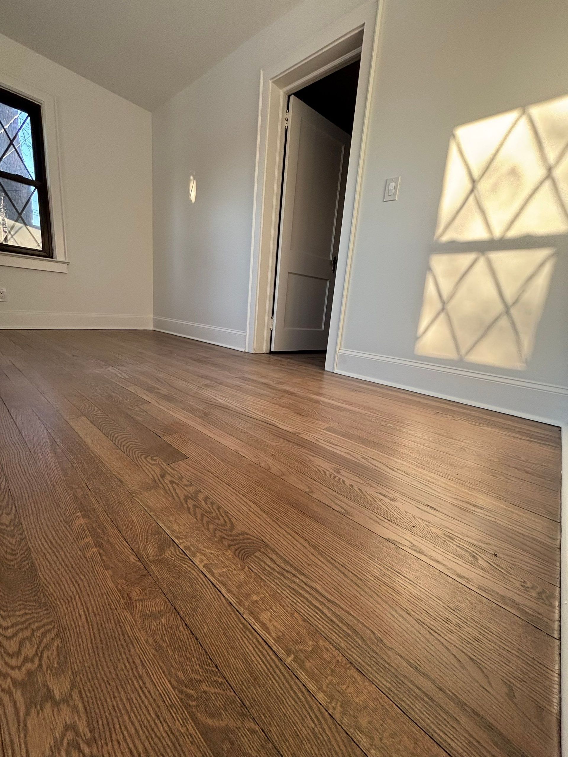 Hardwood floors in a mostly empty room with natural light casting shadows on a wall.