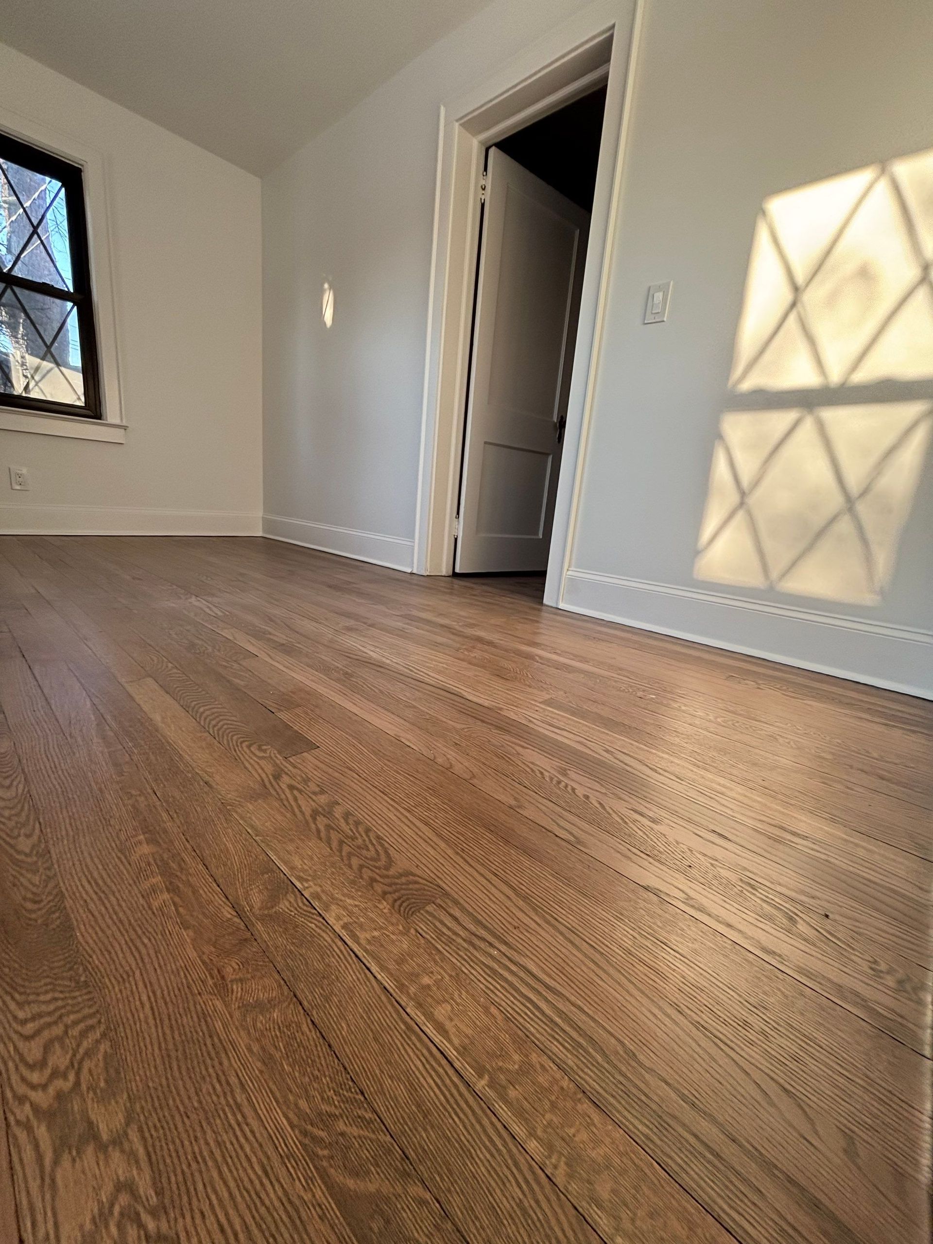 Wooden floor in a room with white walls, door, and window casting a diamond-patterned light.