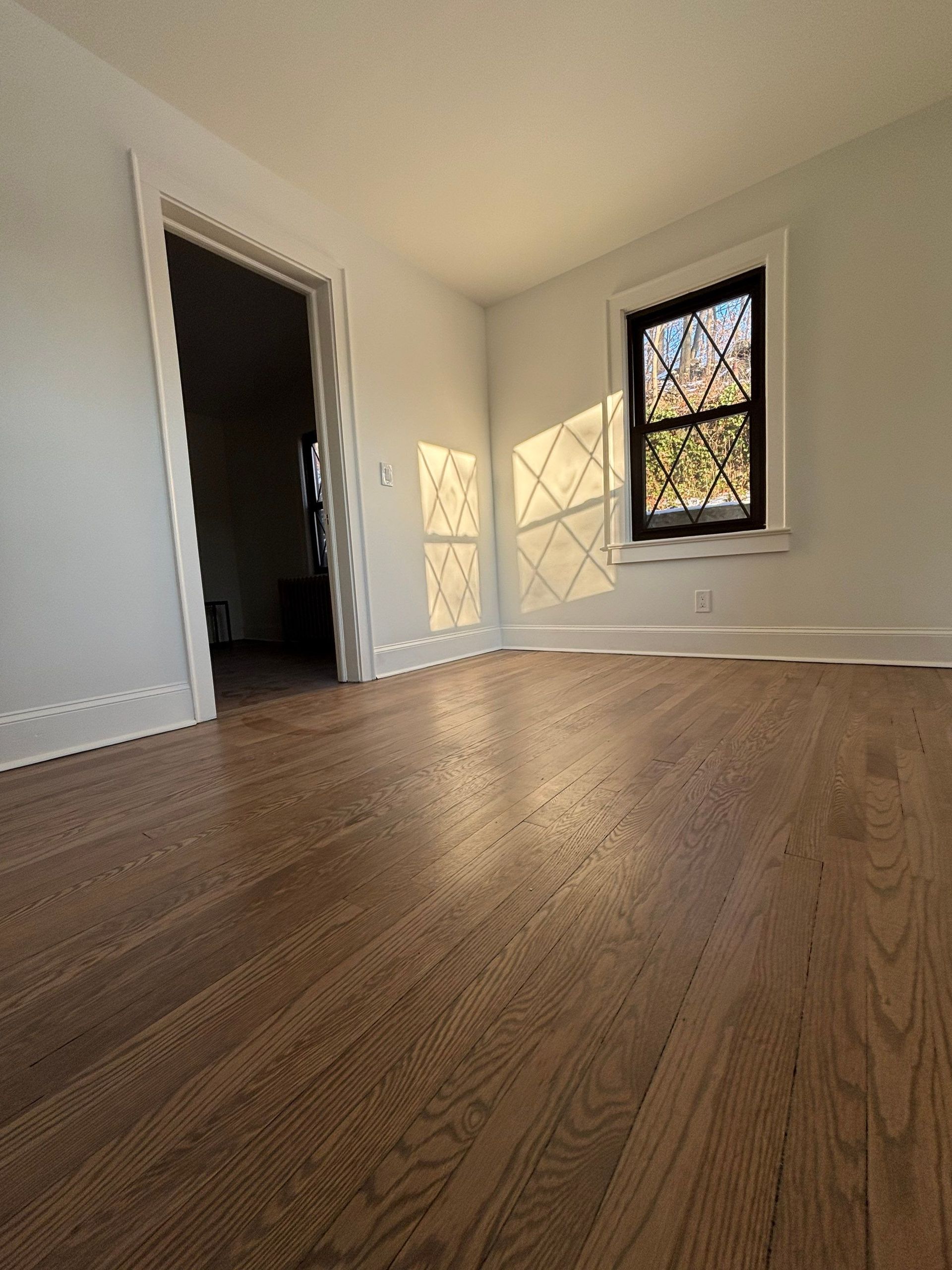 Empty room with hardwood floors, a window, and an open doorway. Sunlight streams across the floor.
