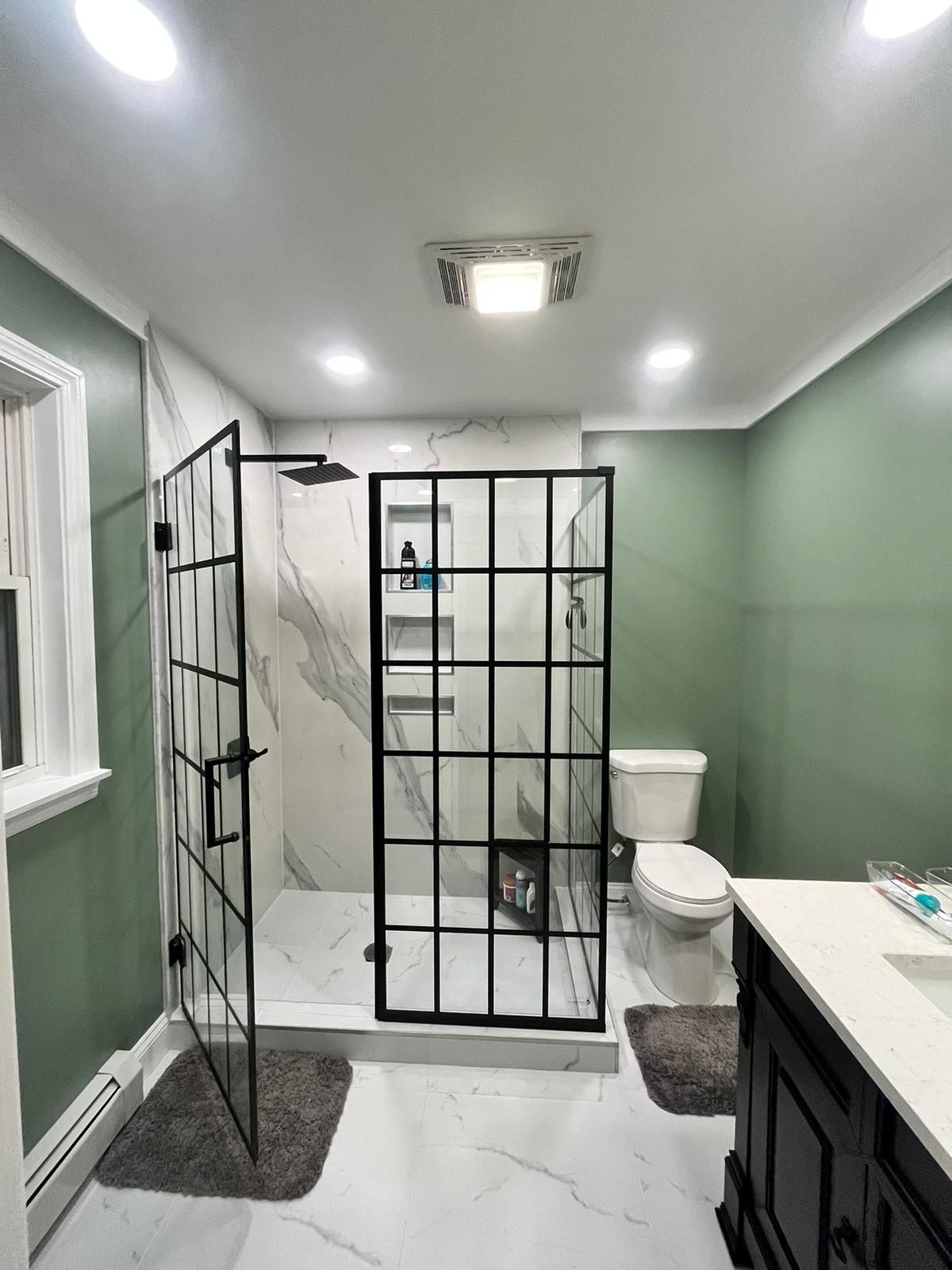 Modern bathroom with green walls, marble shower, black-framed shower doors, toilet, and vanity.