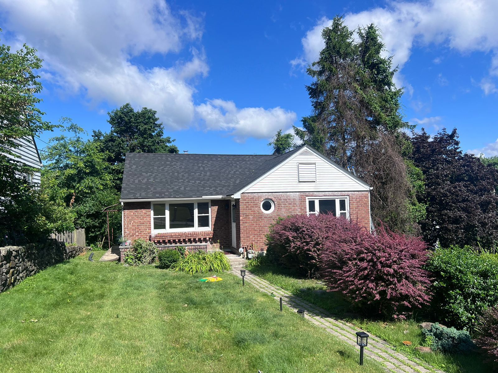 Red brick house with dark roof, surrounded by green grass, bushes, and trees under a blue sky with clouds.