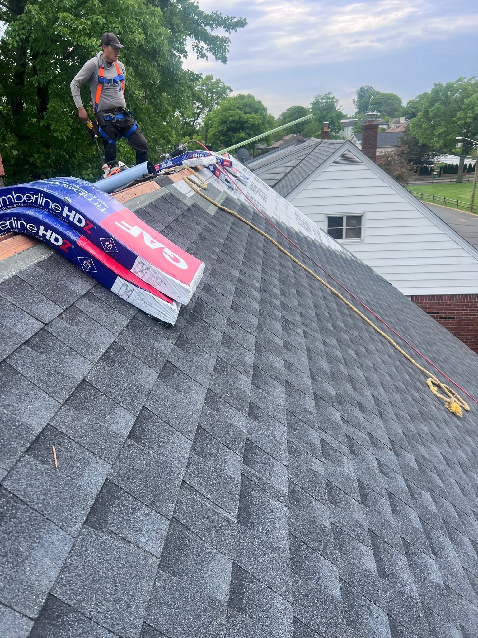 Roofer on a gray shingled roof with safety harness, banners, and rope in an outdoor setting.