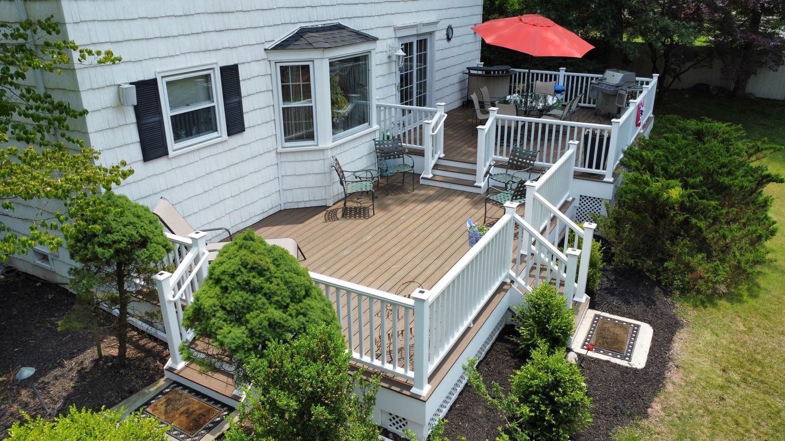 White house with multi-level deck, brown decking, white railings. Red umbrella, chairs, green bushes.