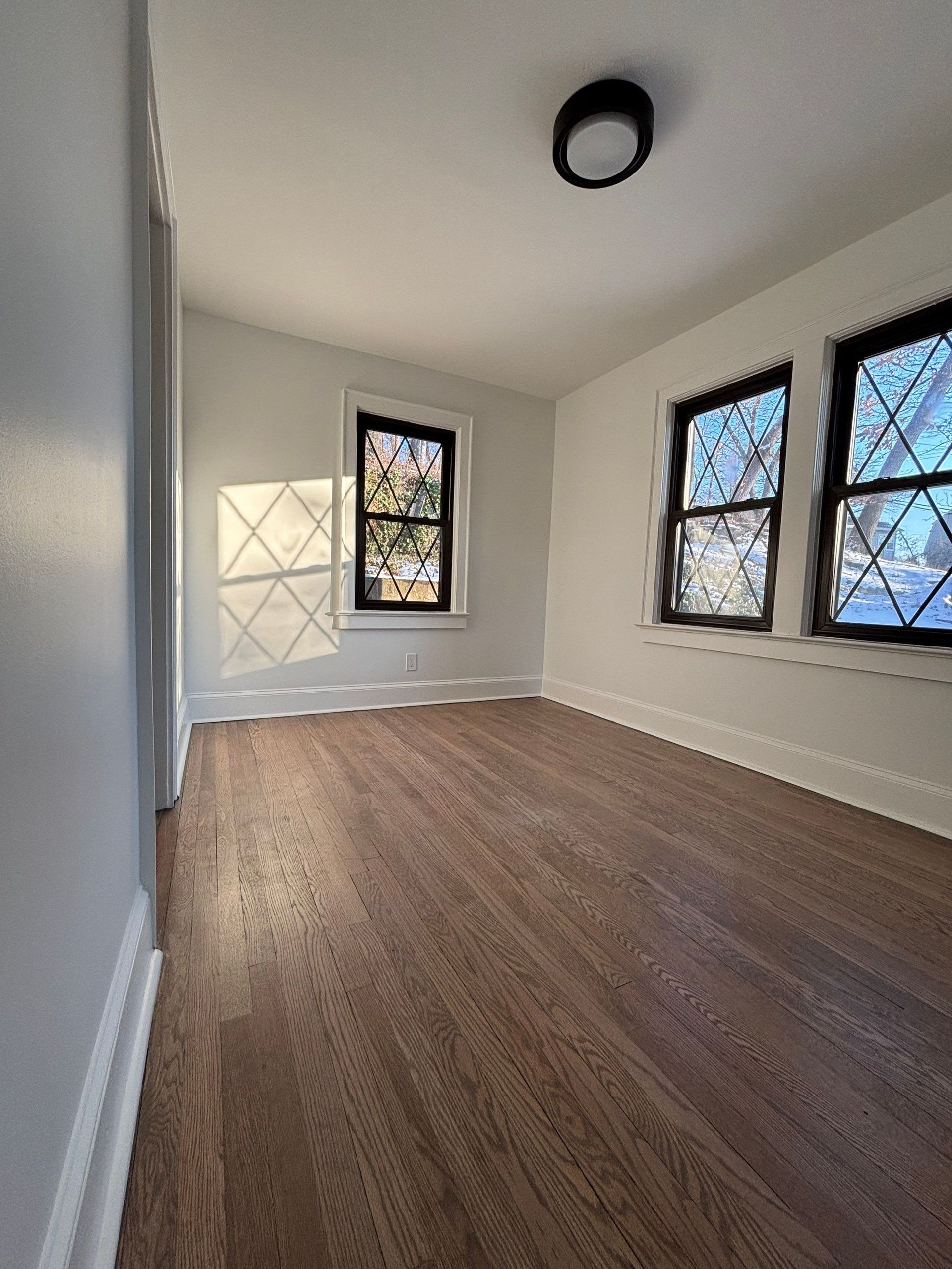 Empty room with hardwood floors, three windows, and a ceiling light. The walls are light blue.