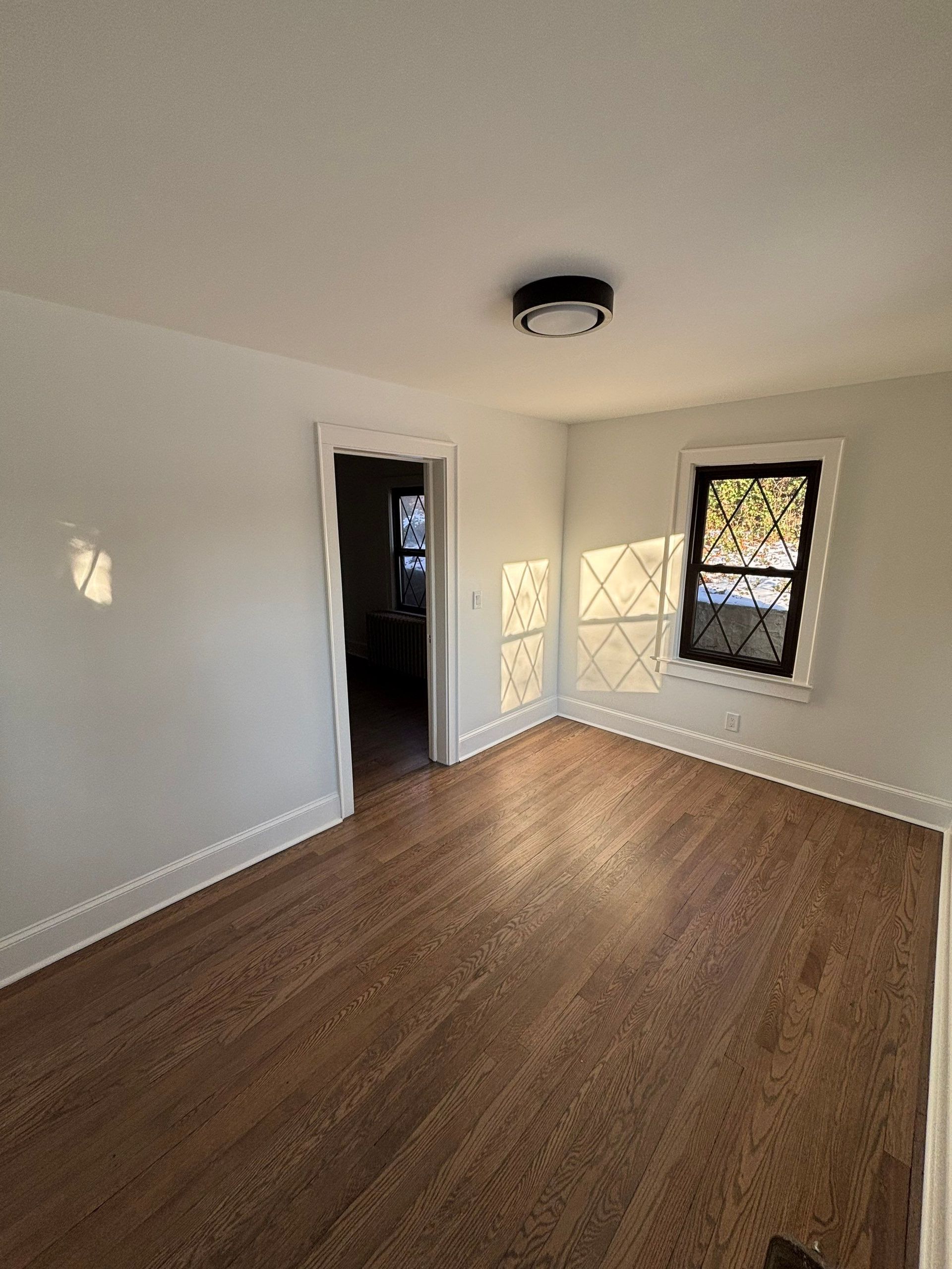 Empty bedroom with hardwood floors, white walls, and doorway. Black-framed window.