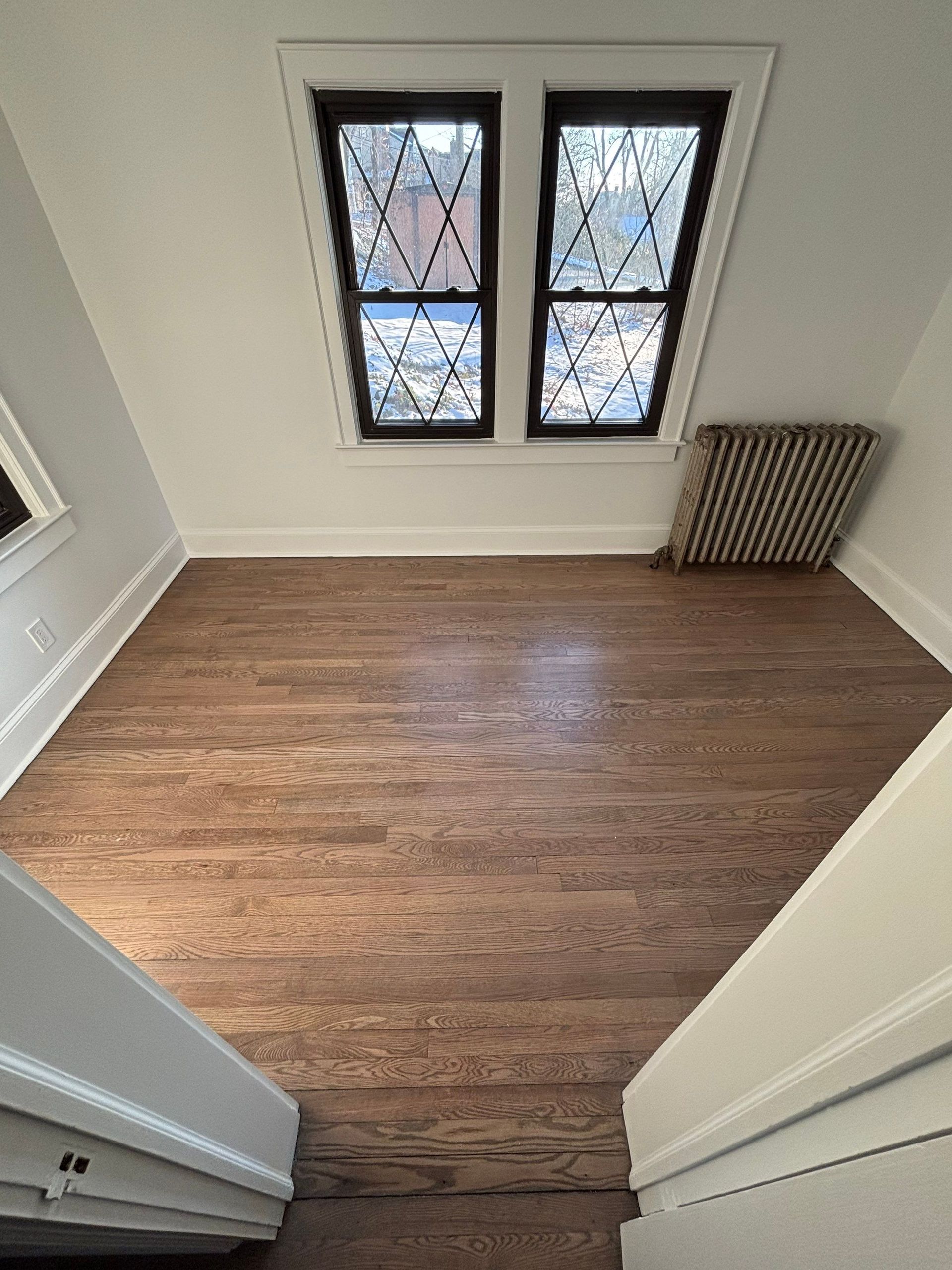 Attic room with hardwood floor, two windows with diamond patterns, radiator, and stairs.
