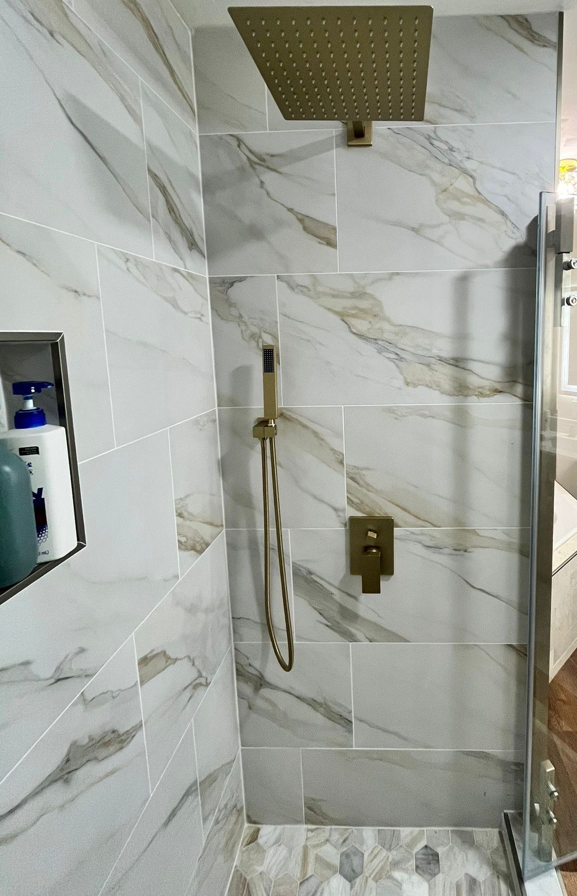 Shower with gold fixtures and marble-patterned tiles, glass door, and a built-in niche for toiletries.