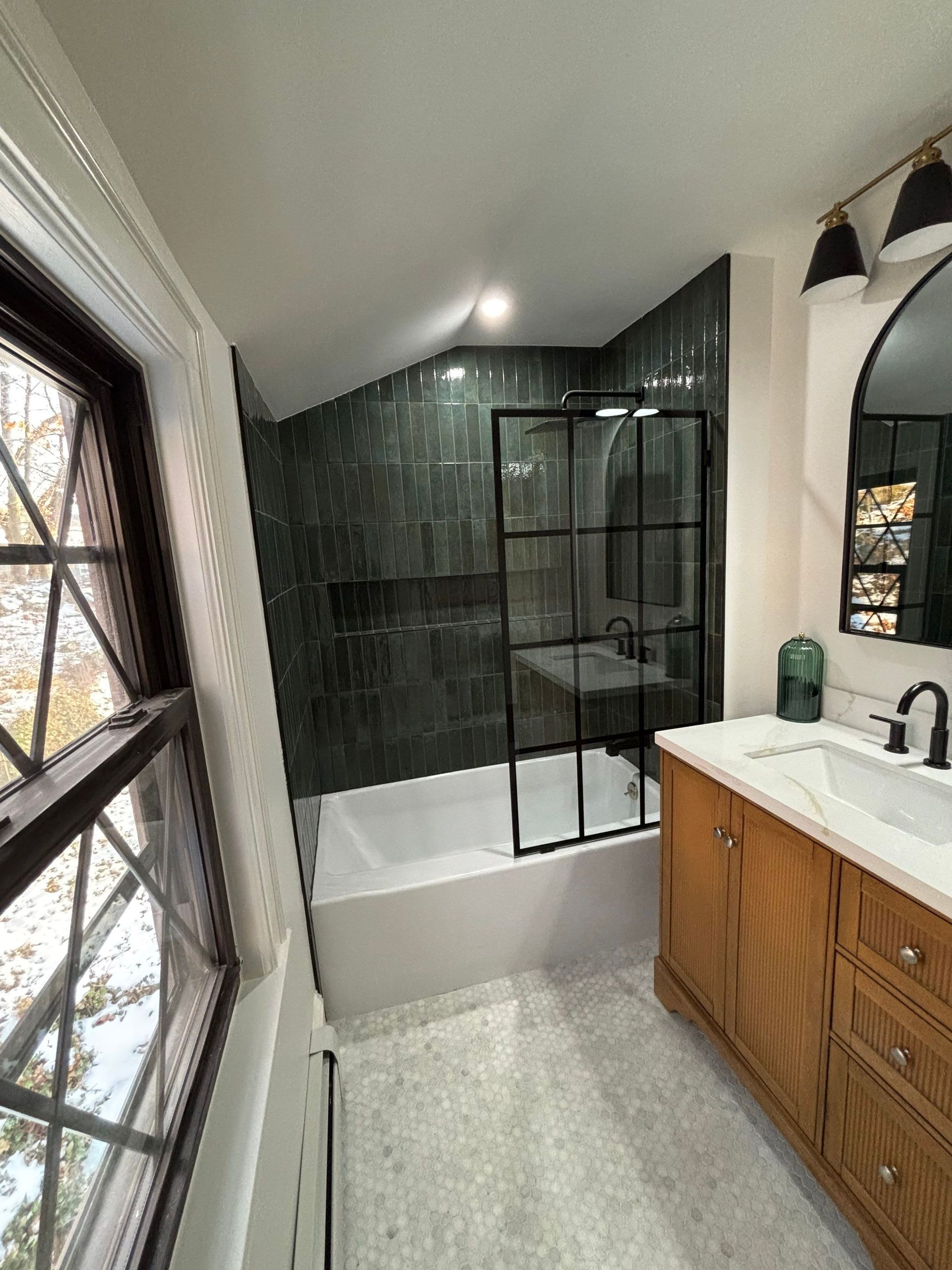 Bathroom with green tiled shower, white tub, wood vanity, and black framed shower door.