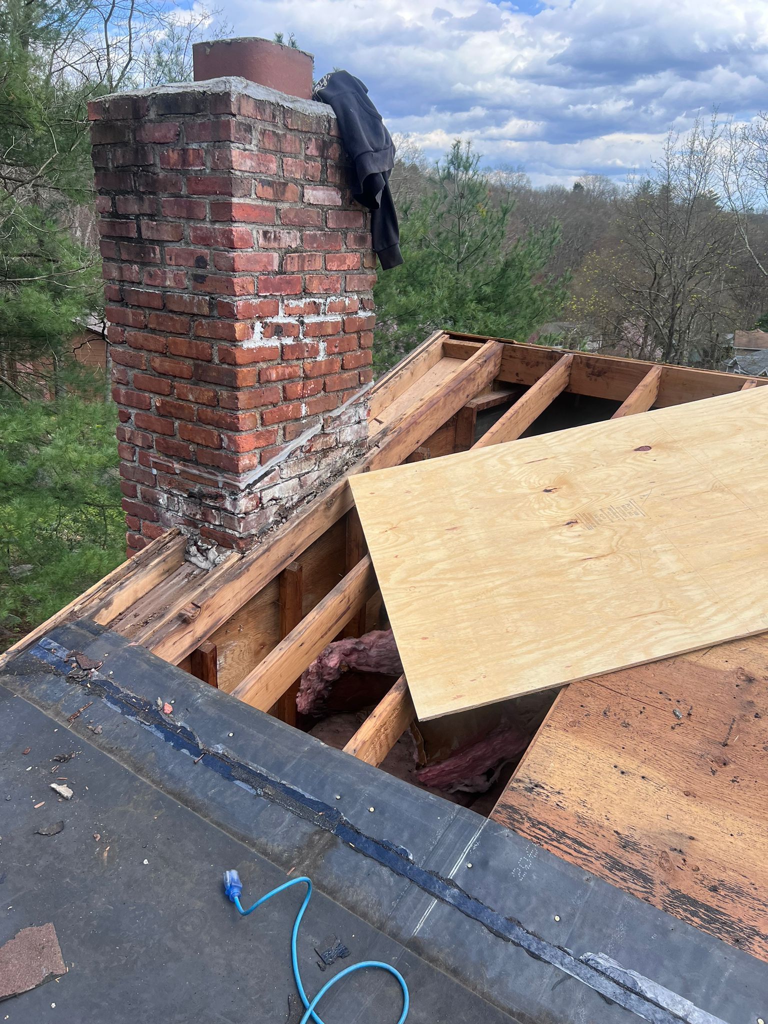 Chimney on a roof with damaged plywood. Brickwork visible. Overcast sky.