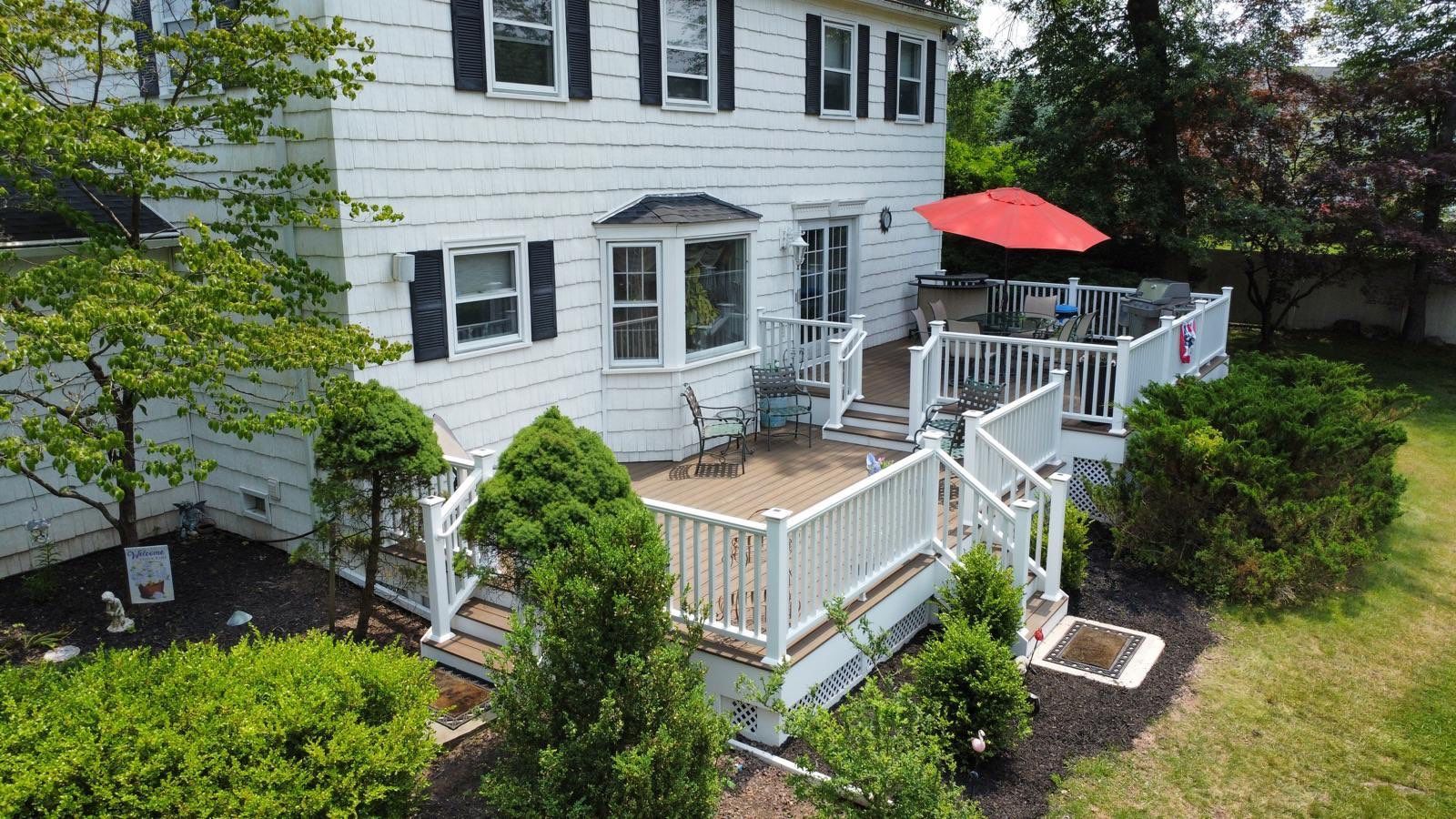 Back of a white house with a multi-level deck, red umbrella, and surrounding greenery.