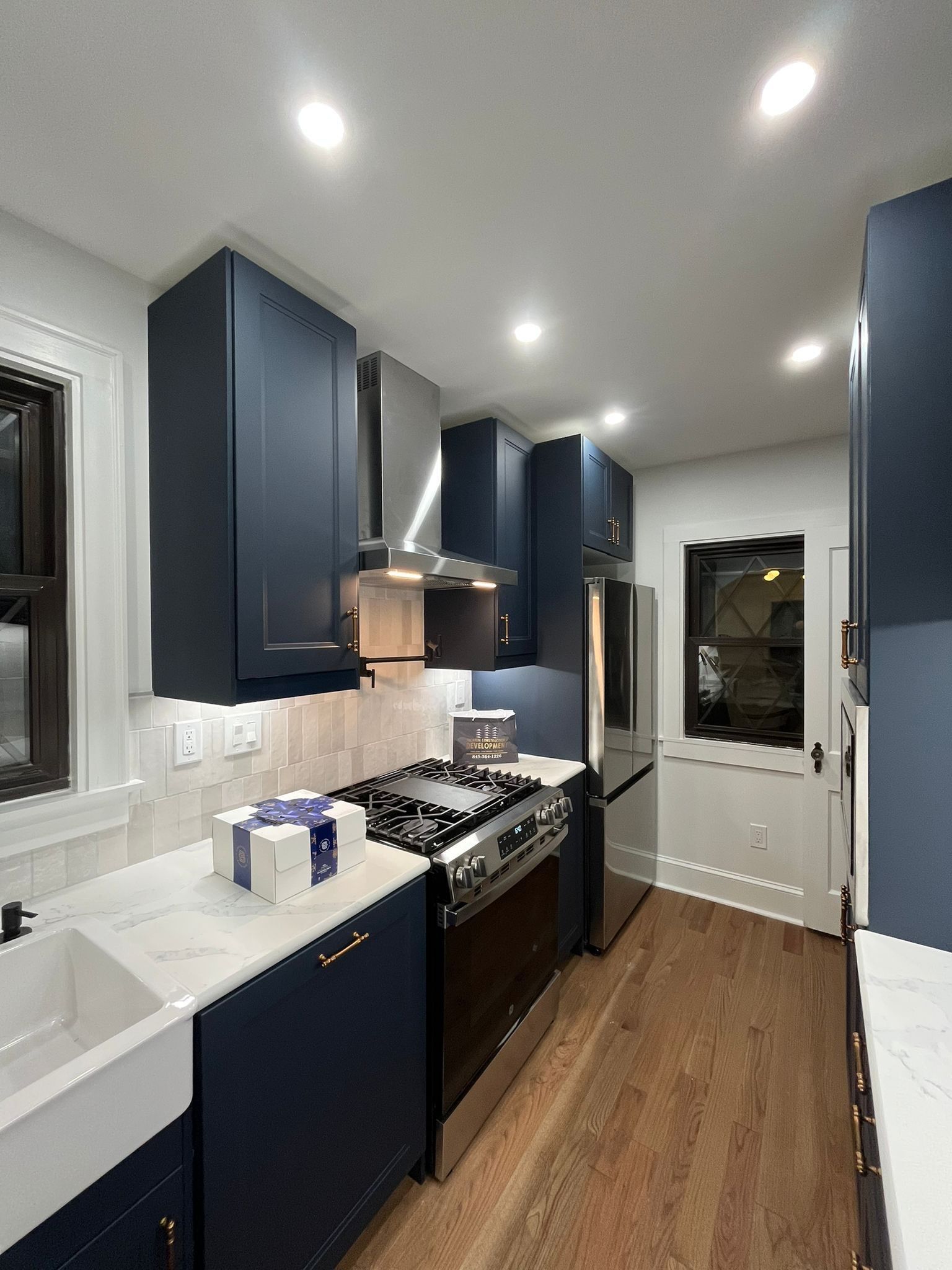 Dark blue kitchen cabinets, stainless steel appliances, white countertops and backsplash, hardwood floor.