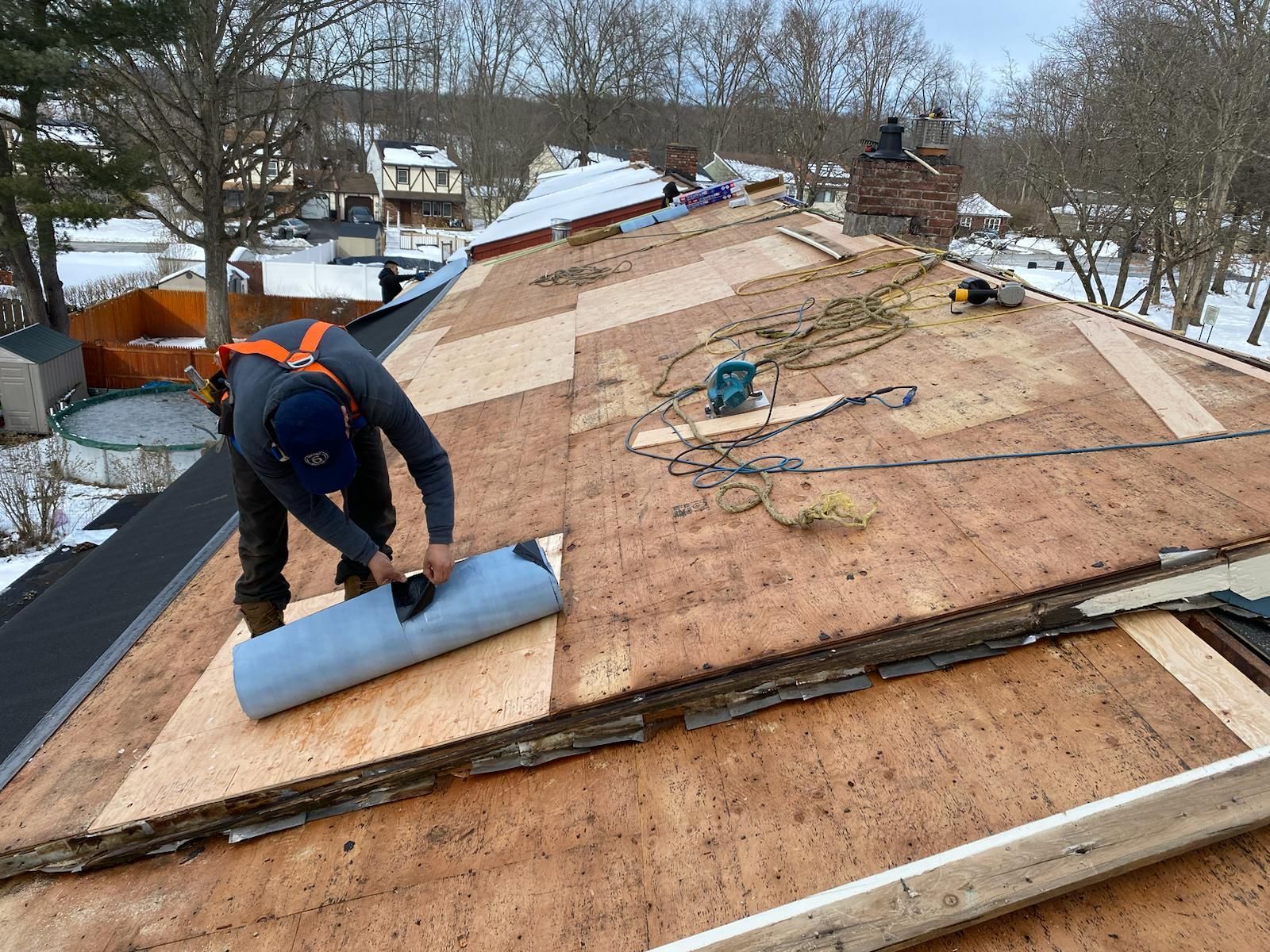 Roofer in safety gear rolls out roofing material on a snowy day.