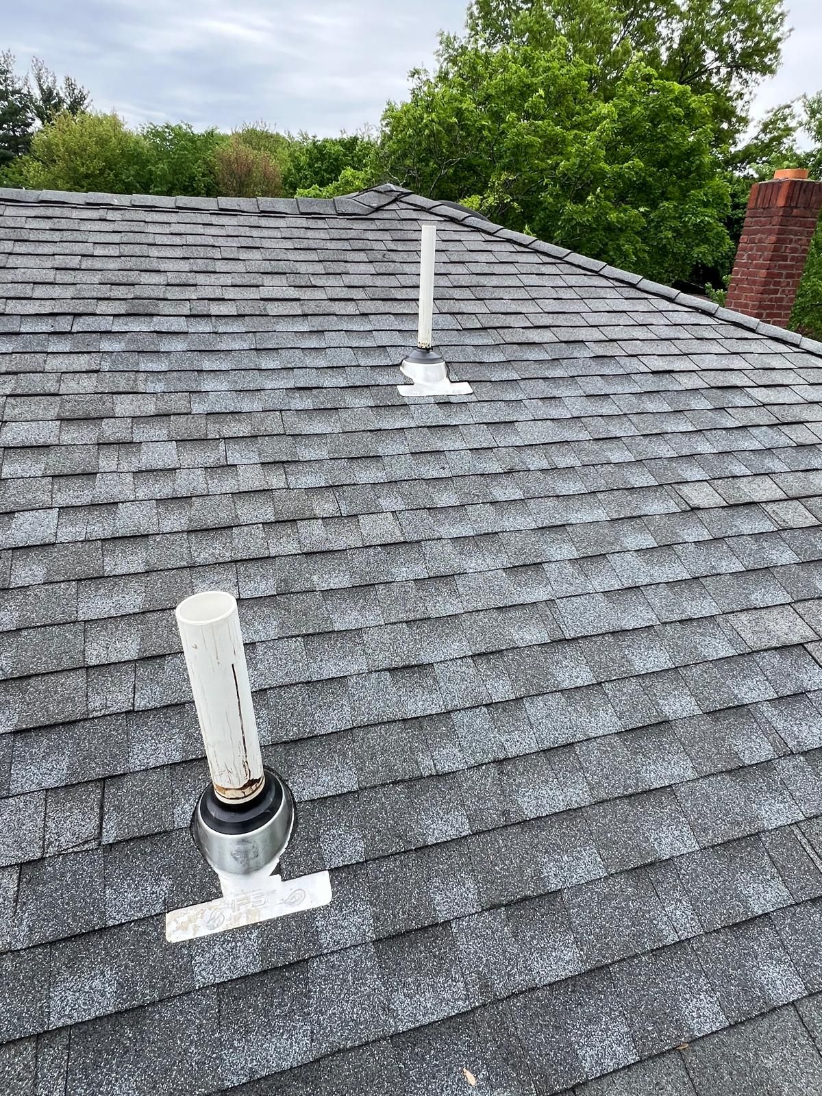 Two white PVC vent pipes on a gray shingle roof. Green trees and a brick chimney are in the background.