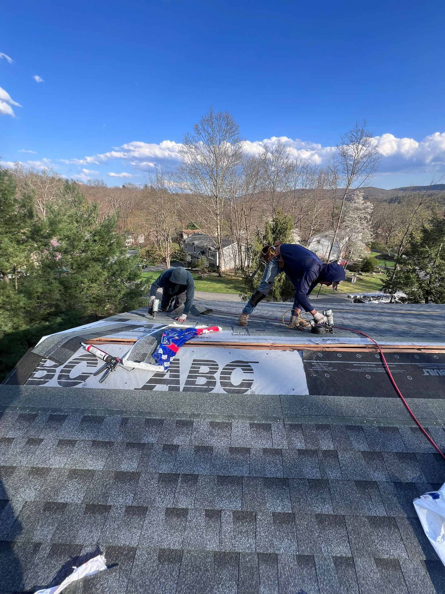 Two roofers working on a shingle roof on a sunny day, trees and a blue sky in the background.