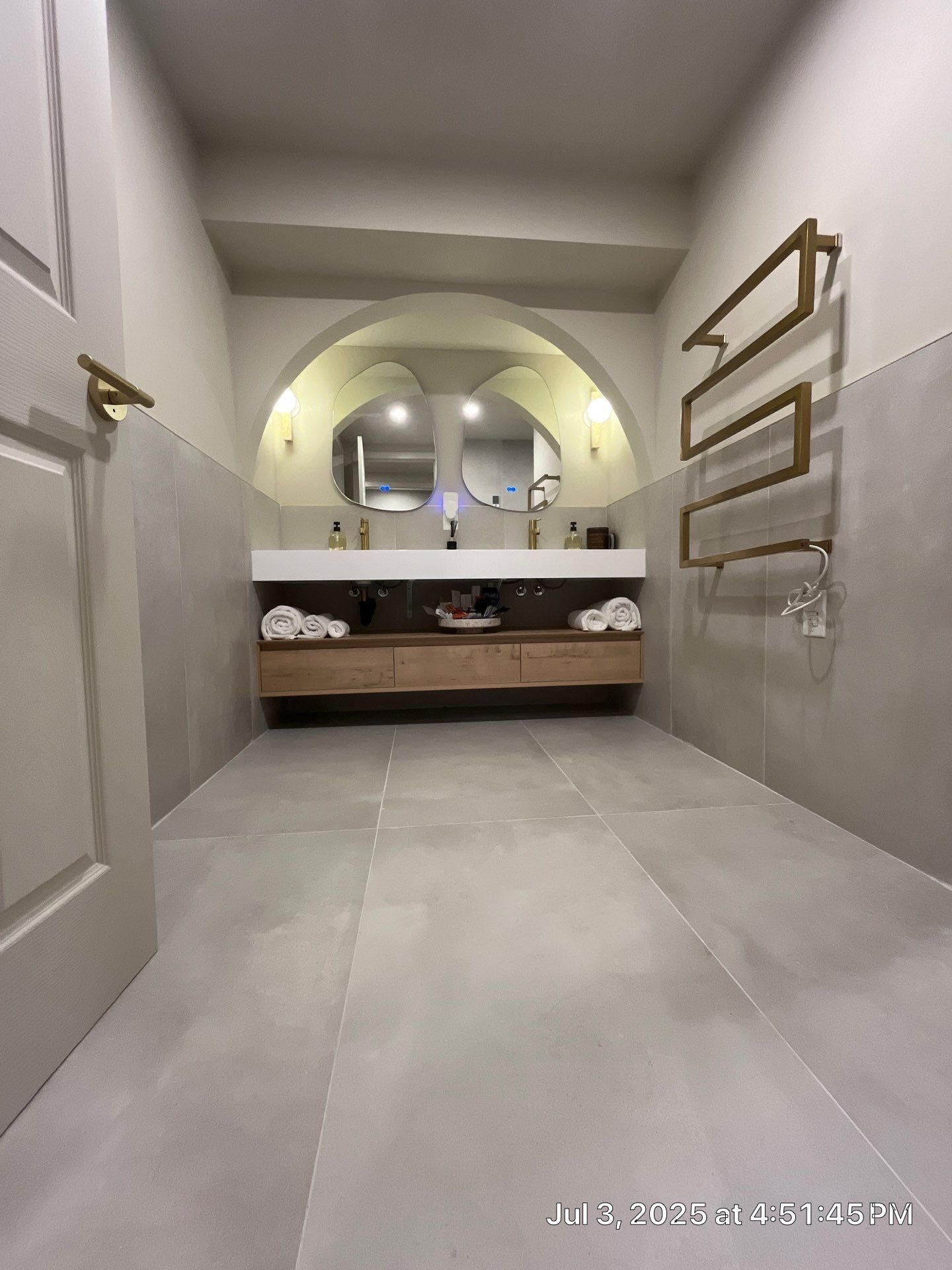 Bathroom with neutral tile, floating wood vanity, arched mirror, and brass towel rack.